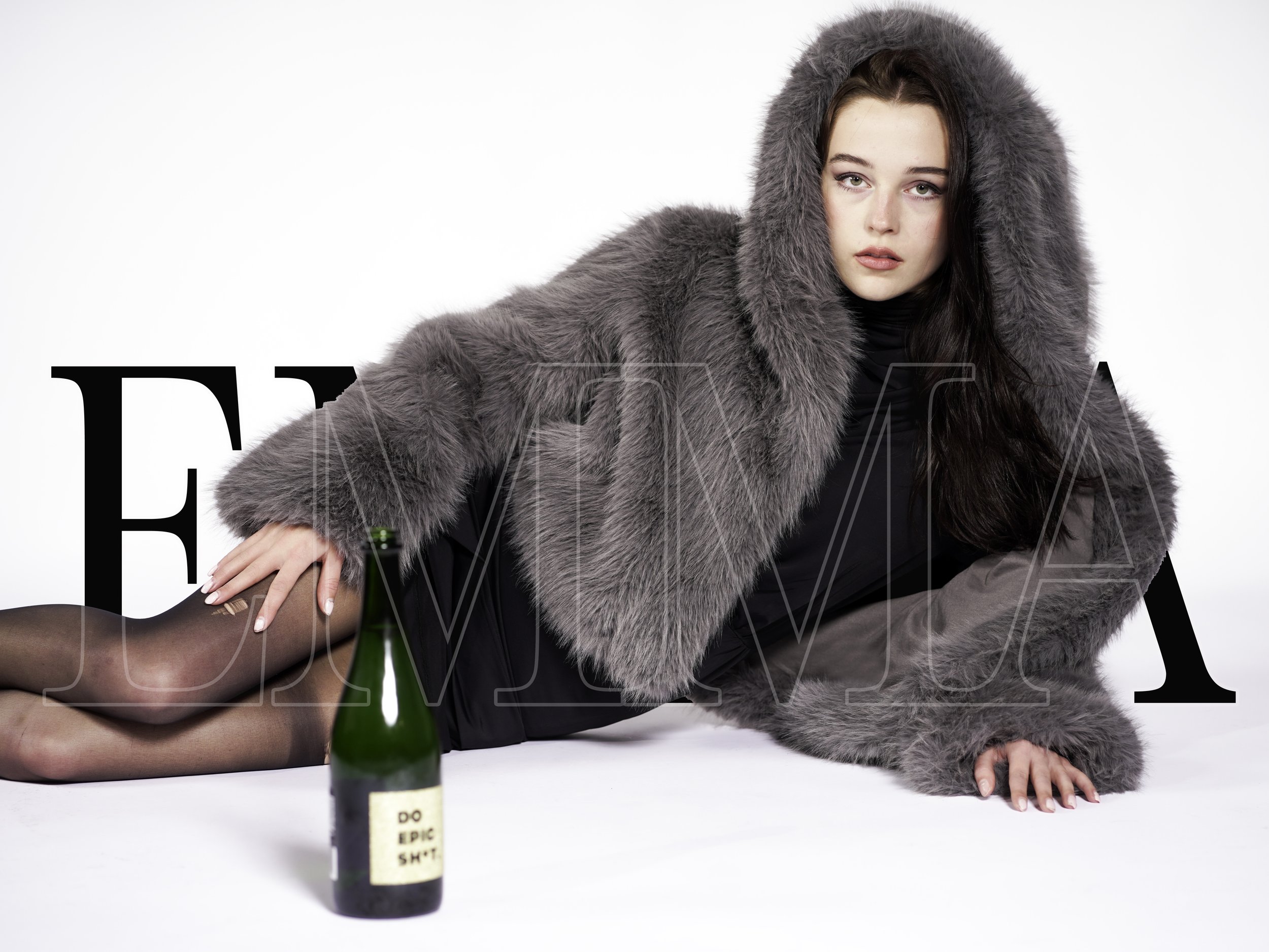 A woman is lying on her side on a white background, wearing a gray fur coat with a hood, a black dress, and black stockings. There is a green bottle with a label that says "DO EPIC SHIT" in front of her.