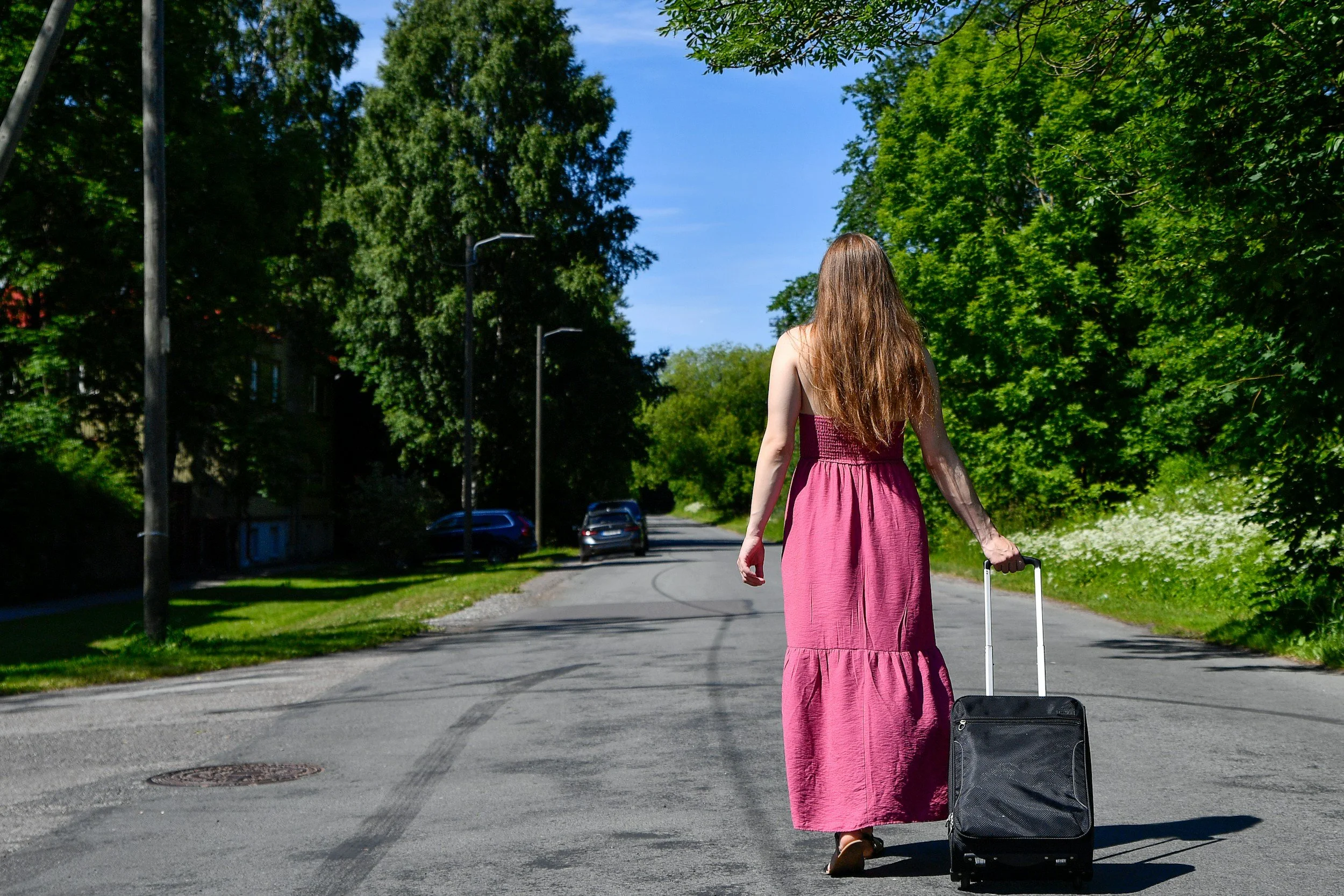 A woman with long brown hair wearing a sleeveless pink dress walking down a suburban street pulling a small black rolling suitcase, with trees and parked cars visible along the street.