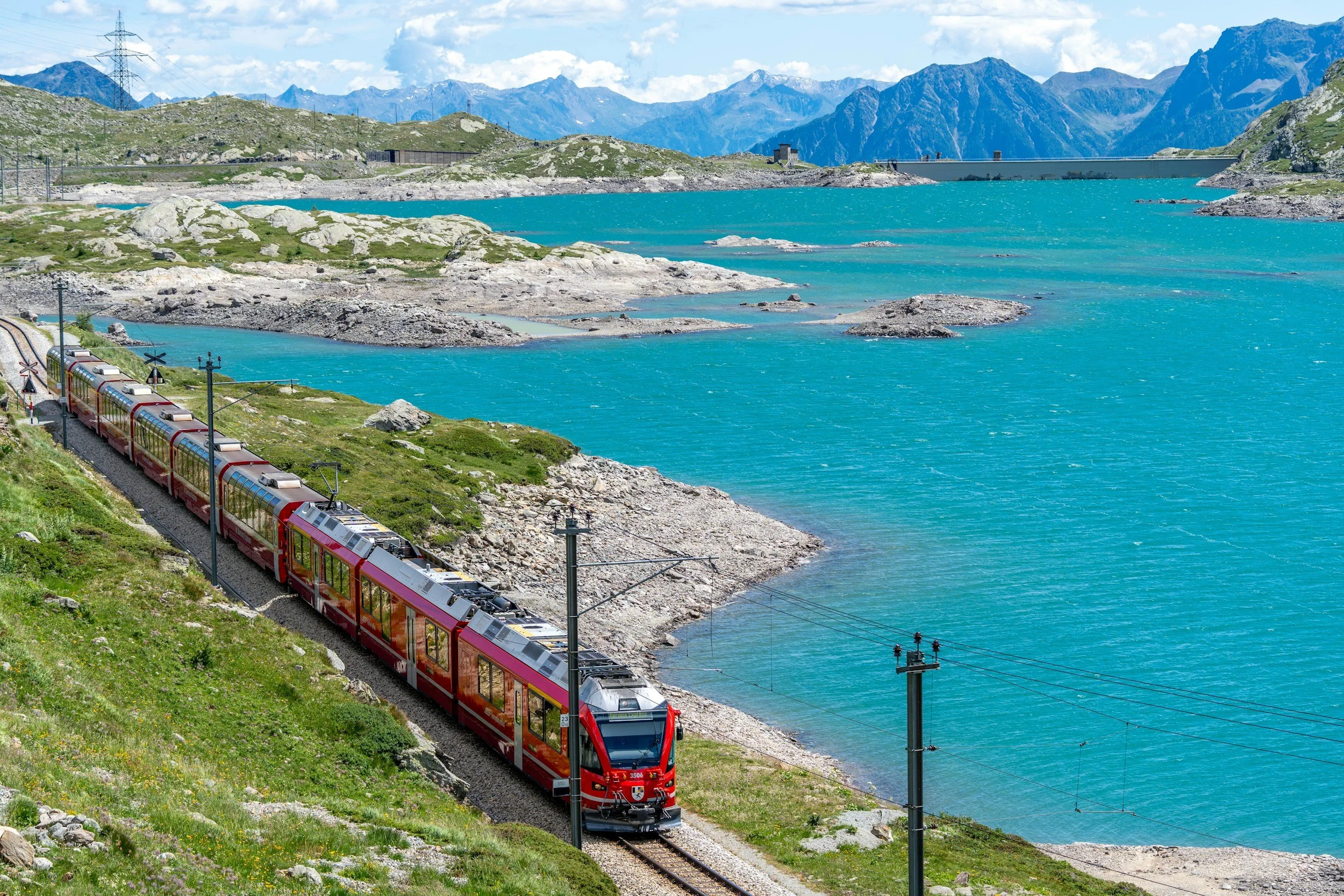 A red passenger train traveling along a railway track beside a turquoise lake with mountainous terrain and a partly cloudy sky in the background.