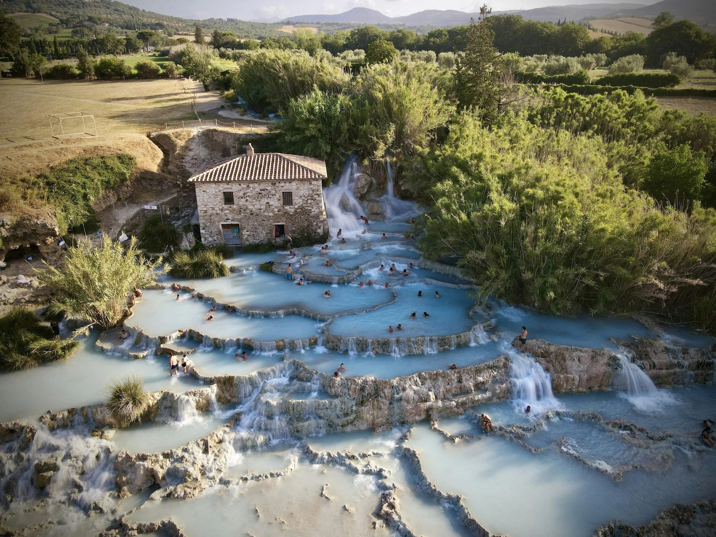 People swimming and relaxing in natural hot springs with cascading waterfalls surrounded by lush greenery and mountains.