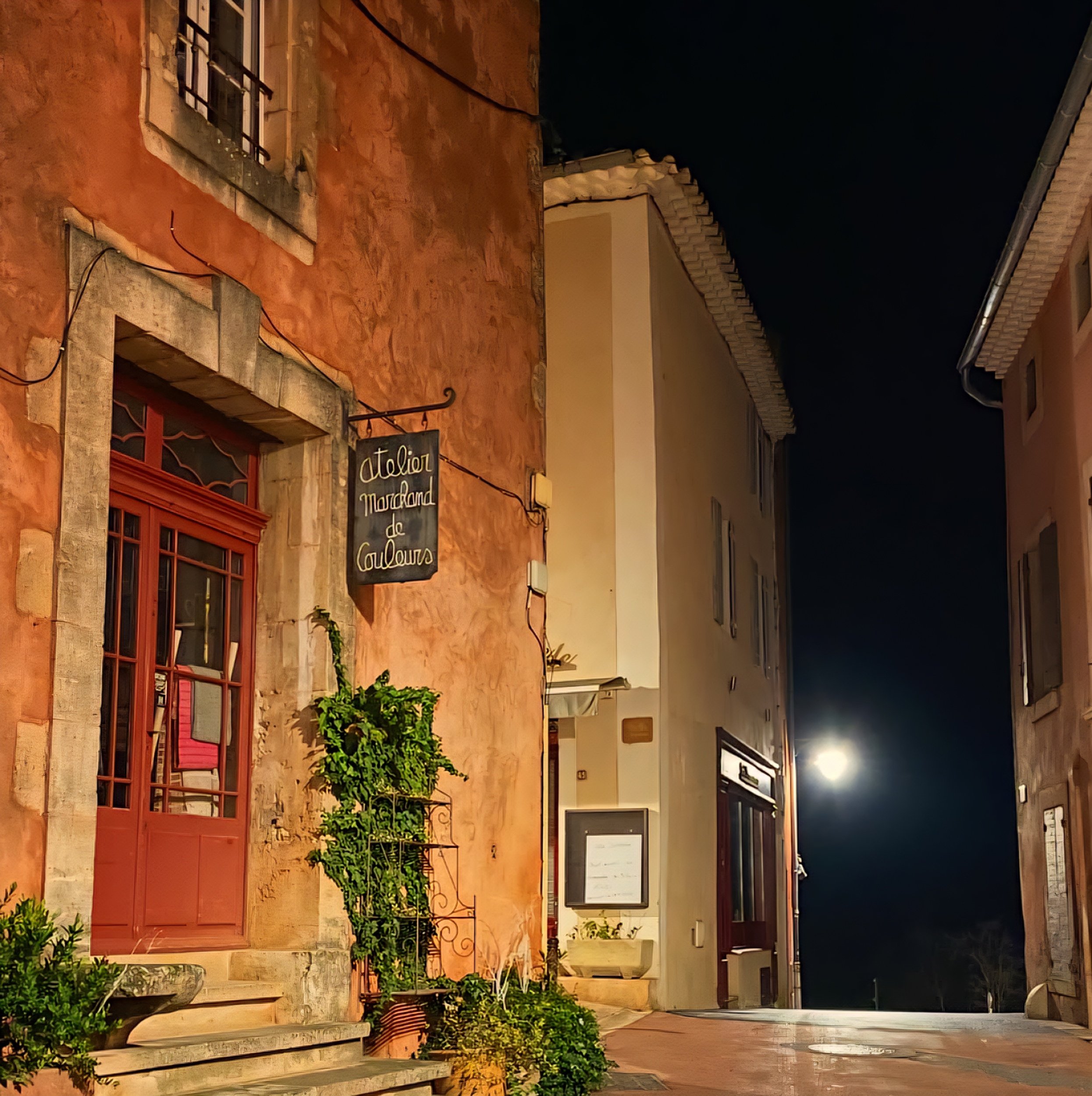 Night view of a narrow street with buildings; one building has a sign that reads 'Atelier Mourand de Couleurs' and features a window with red shutters and potted plants, illuminated by streetlights.
