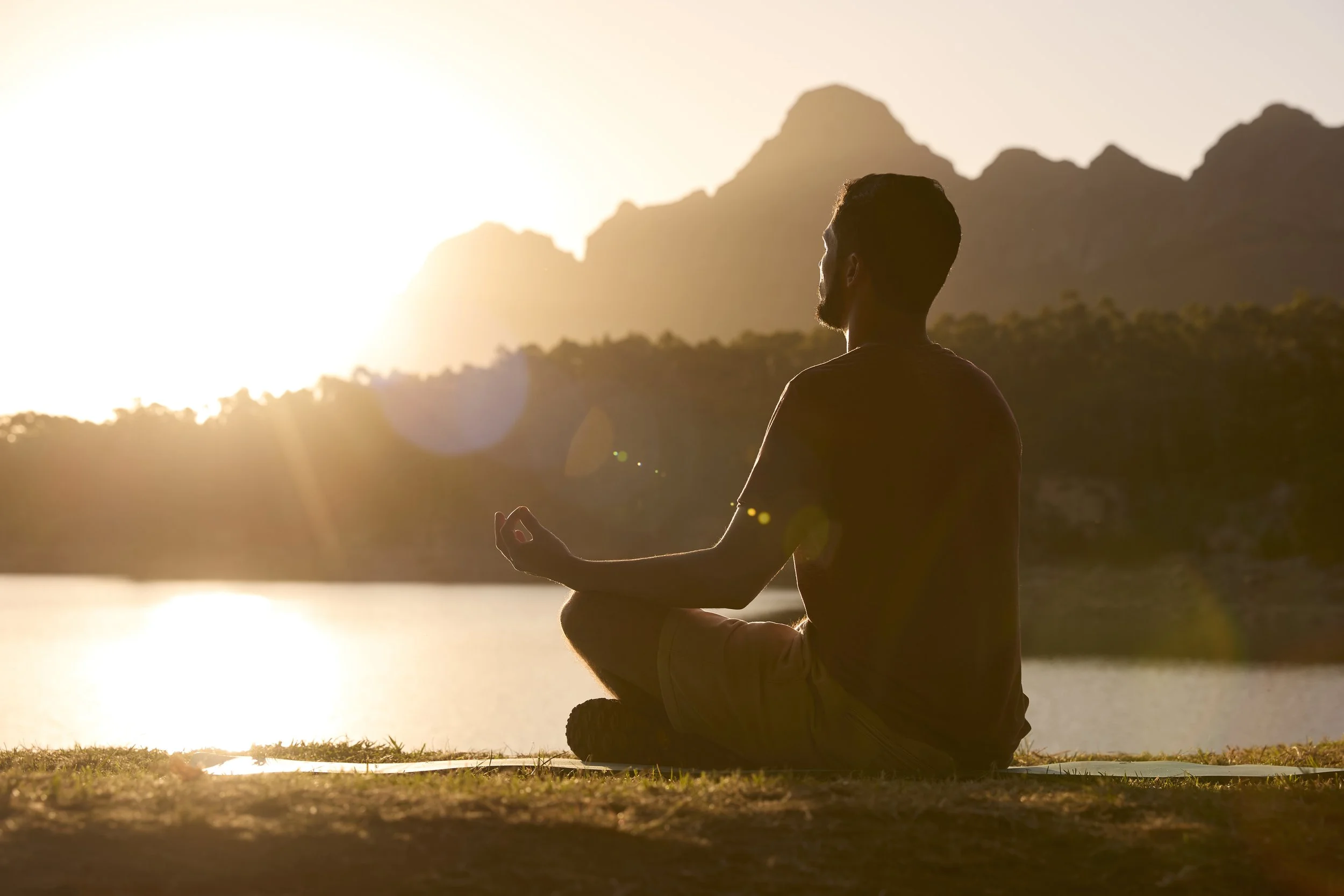 A person practicing yoga or meditation by a lake during sunset, with mountains in the background.