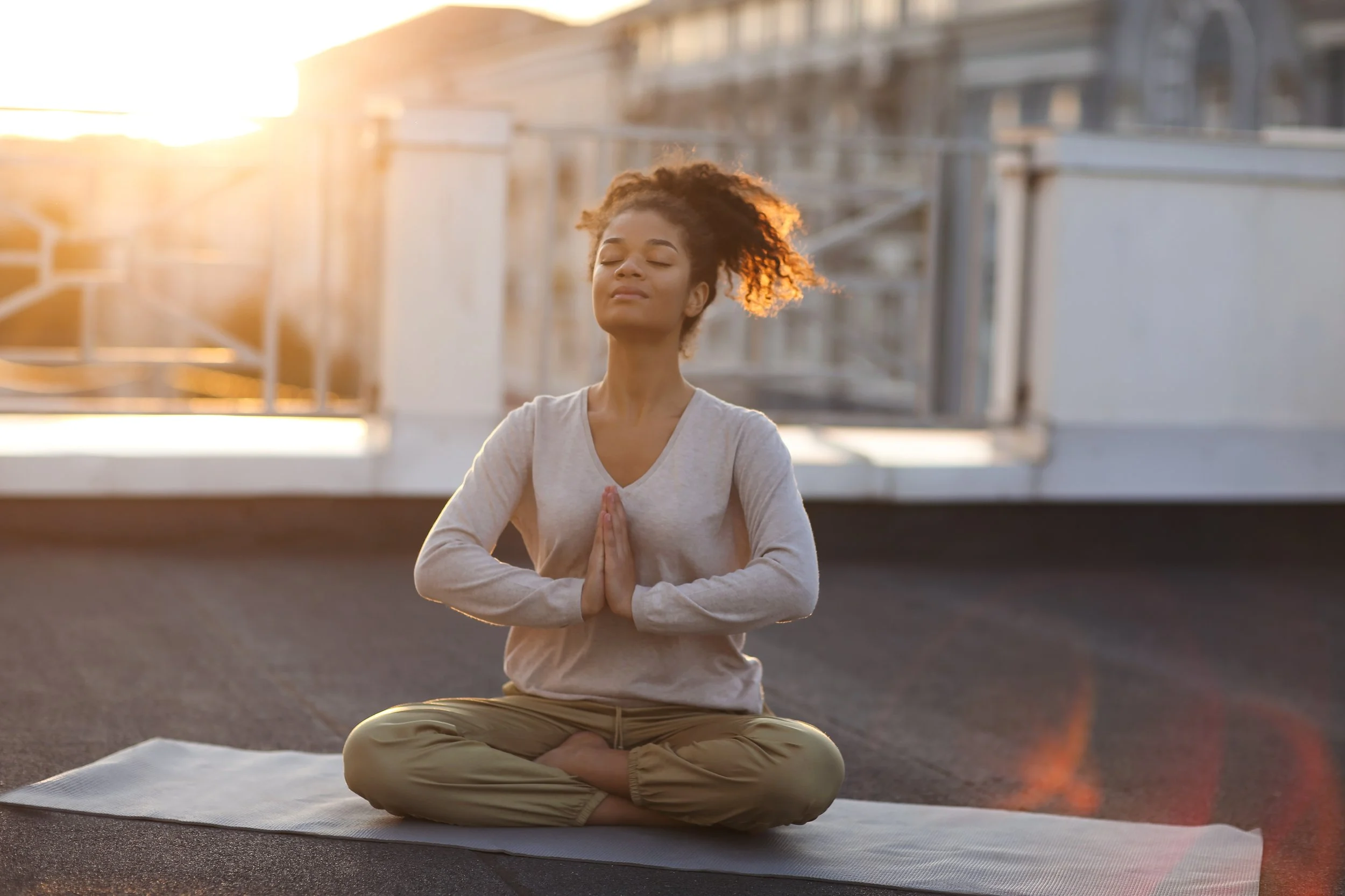 A woman practicing yoga or meditation outdoors on a mat at sunset, sitting cross-legged with hands in prayer position, eyes closed, and a serene expression.