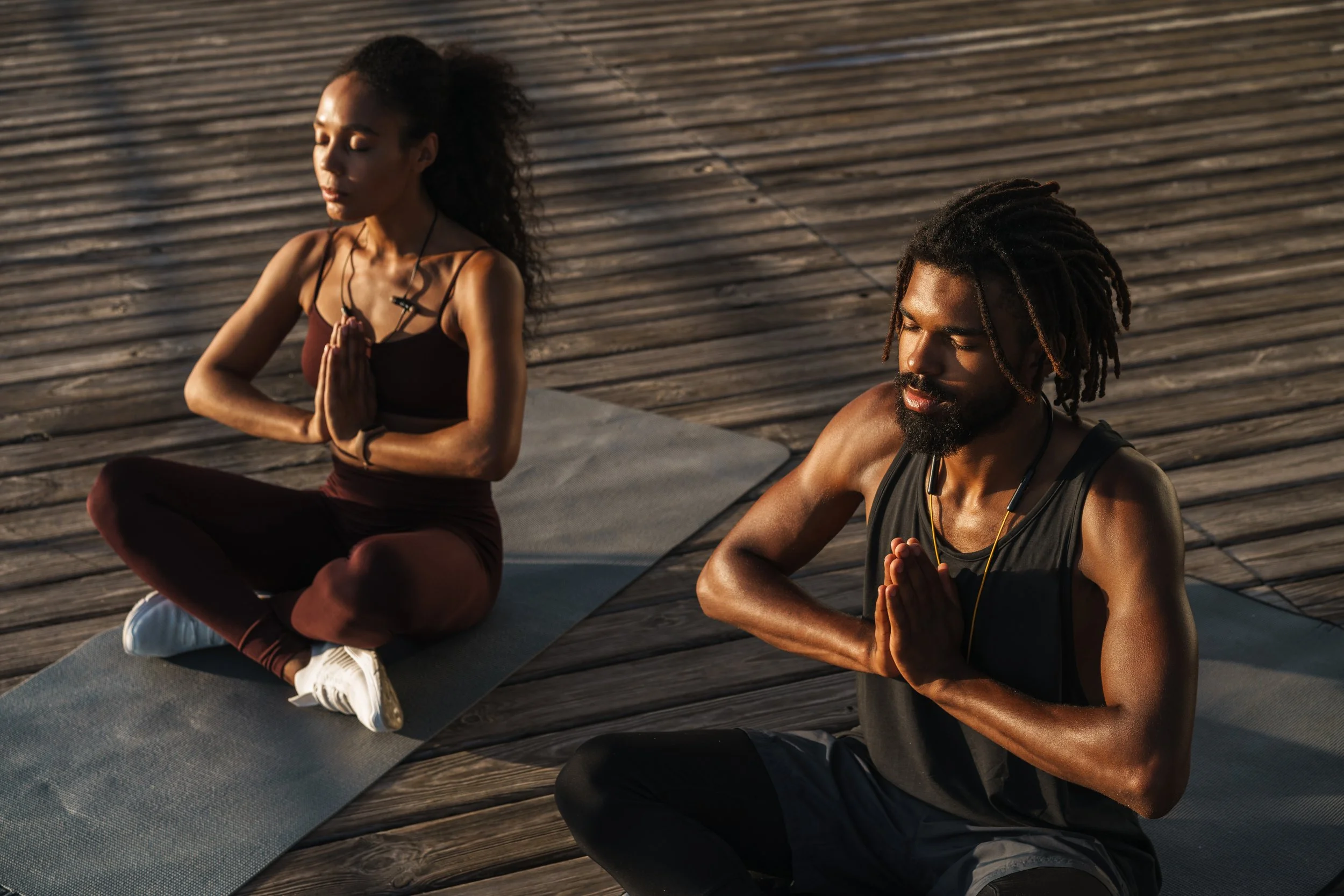 Two individuals practicing yoga outdoors on a wooden deck during sunset, sitting cross-legged with hands in prayer position, eyes closed, wearing athletic clothing and earphones.