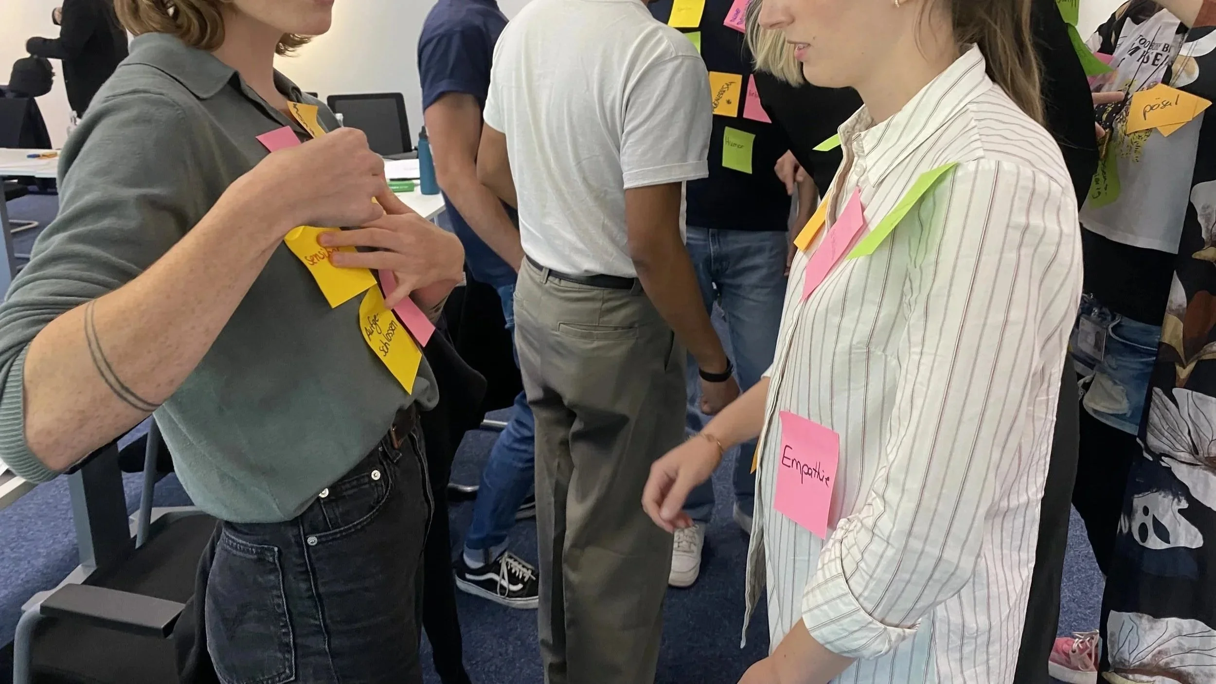 Two women engaging in a conversation at a workshop or event, with multiple colorful sticky notes attached to their clothing and surrounding participants.