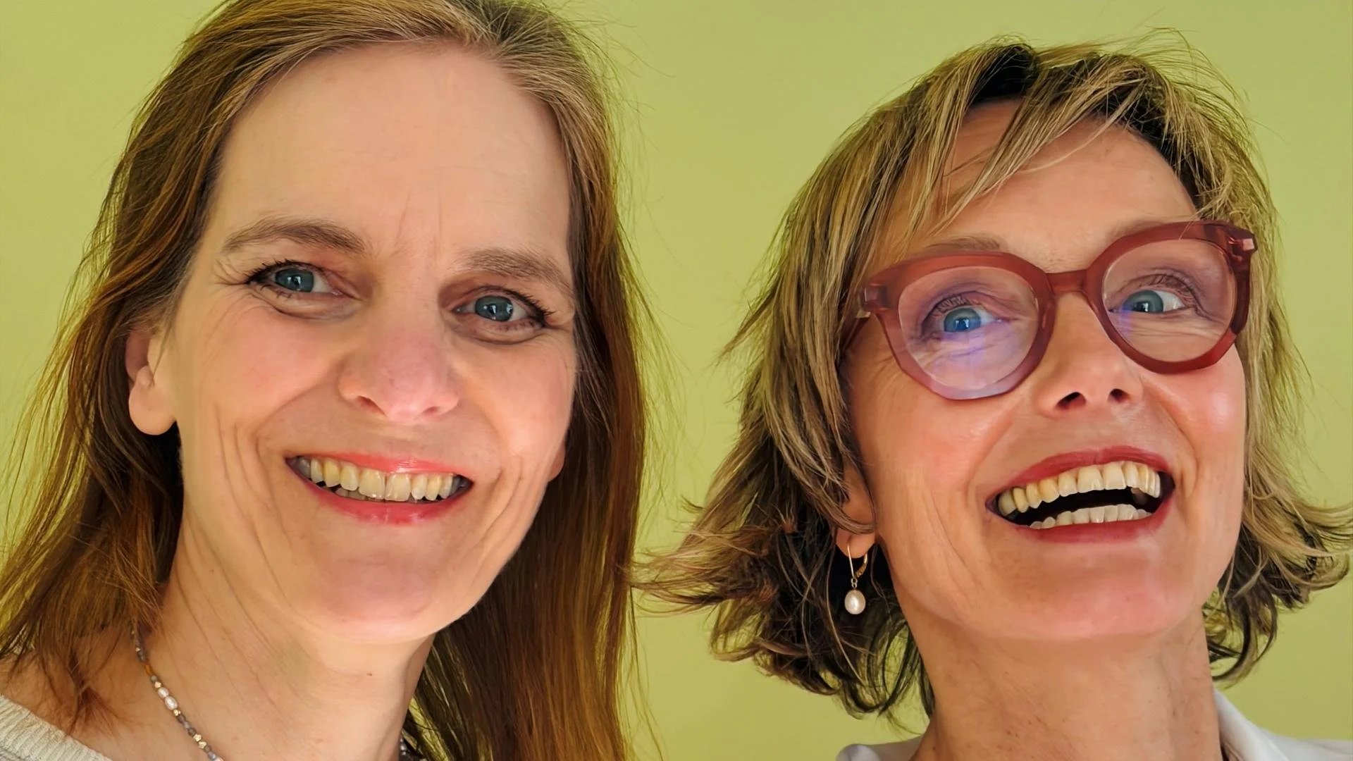 Close-up of two women smiling, one with red hair and the other with dark blonde hair, wearing glasses and earrings, against a light yellow background.
