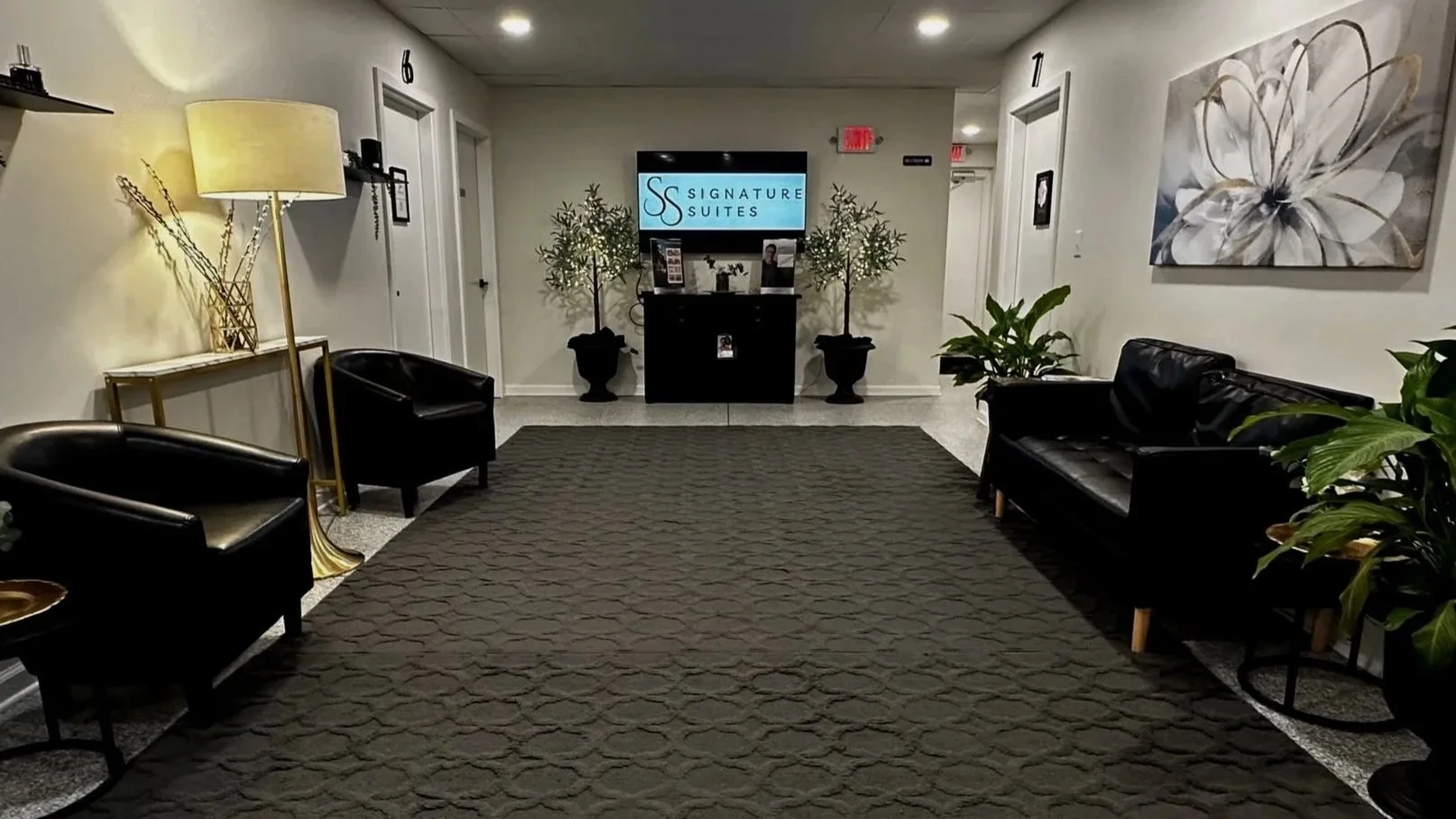 Suite lobby with black leather chairs and a patterned carpet, wall art, potted plants, a large flat screen displaying 'Signature Suites,' and a small console table with a lamp.