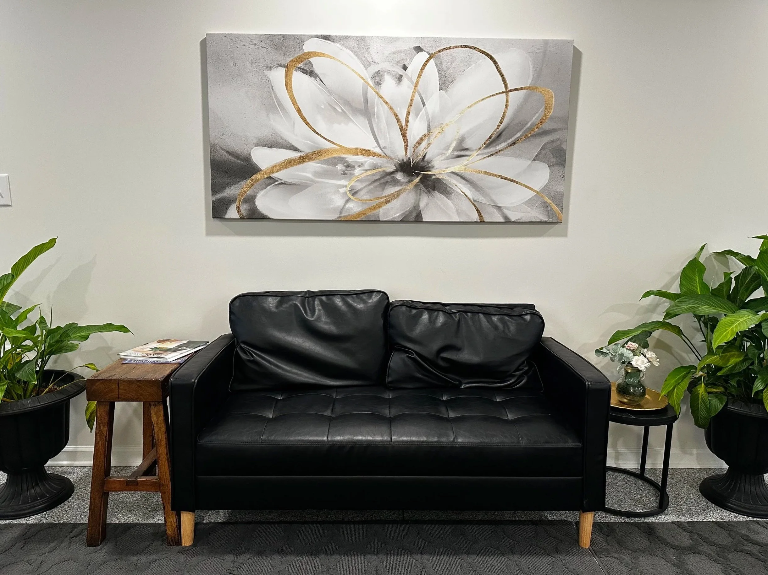 Suite lobby with a black leather loveseat, a wooden side table, and two black planters with green plants on either side. A large gray and white abstract flower painting with gold accents hangs on the wall behind the loveseat.
