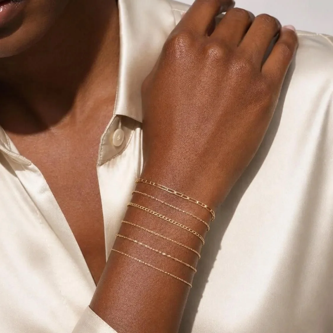 A person wearing multiple delicate gold bracelets on their wrist, dressed in a cream-colored satin blouse.
