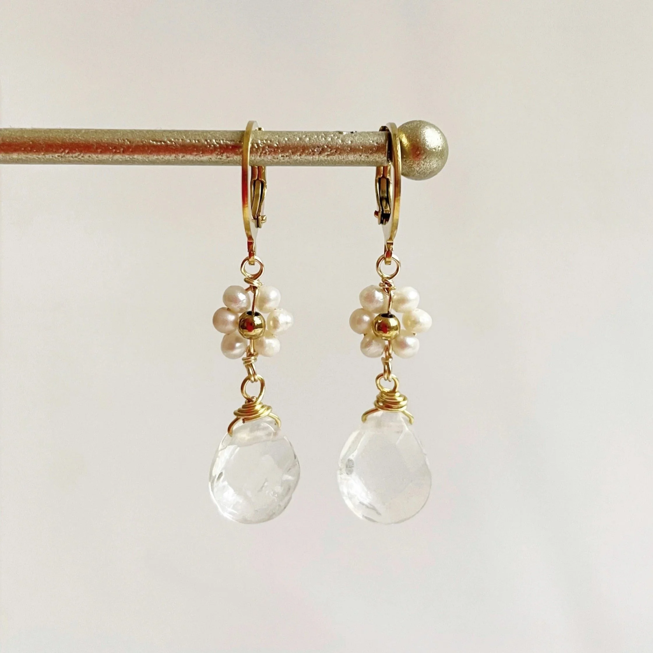 A jewelry display with gold and pearl earrings hanging from a horizontal metal rod against a plain white background.