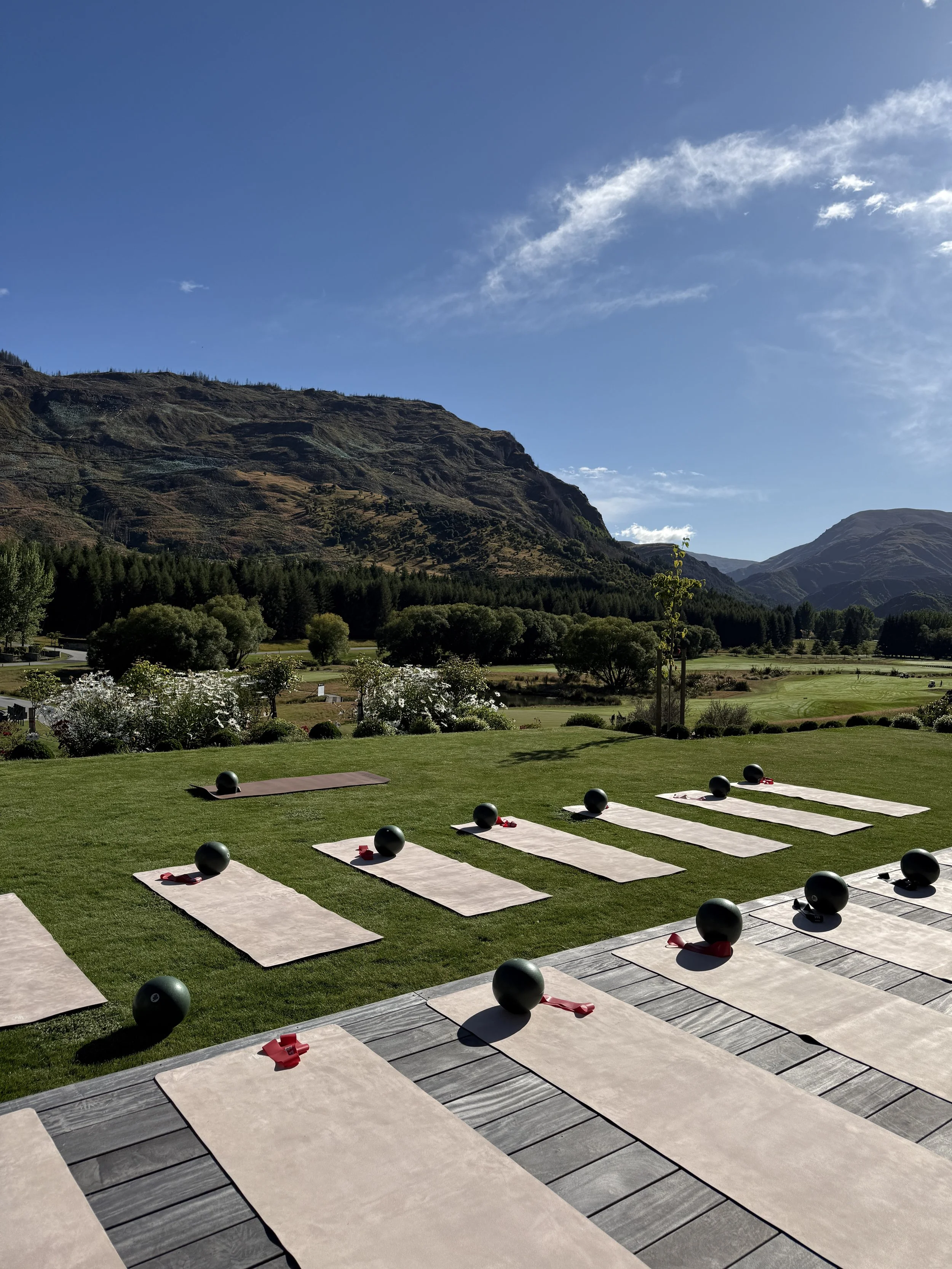 Outdoor yoga or meditation setup with mats, black spheres, and red resistance bands on a grassy area with mountains and blue sky in the background.
