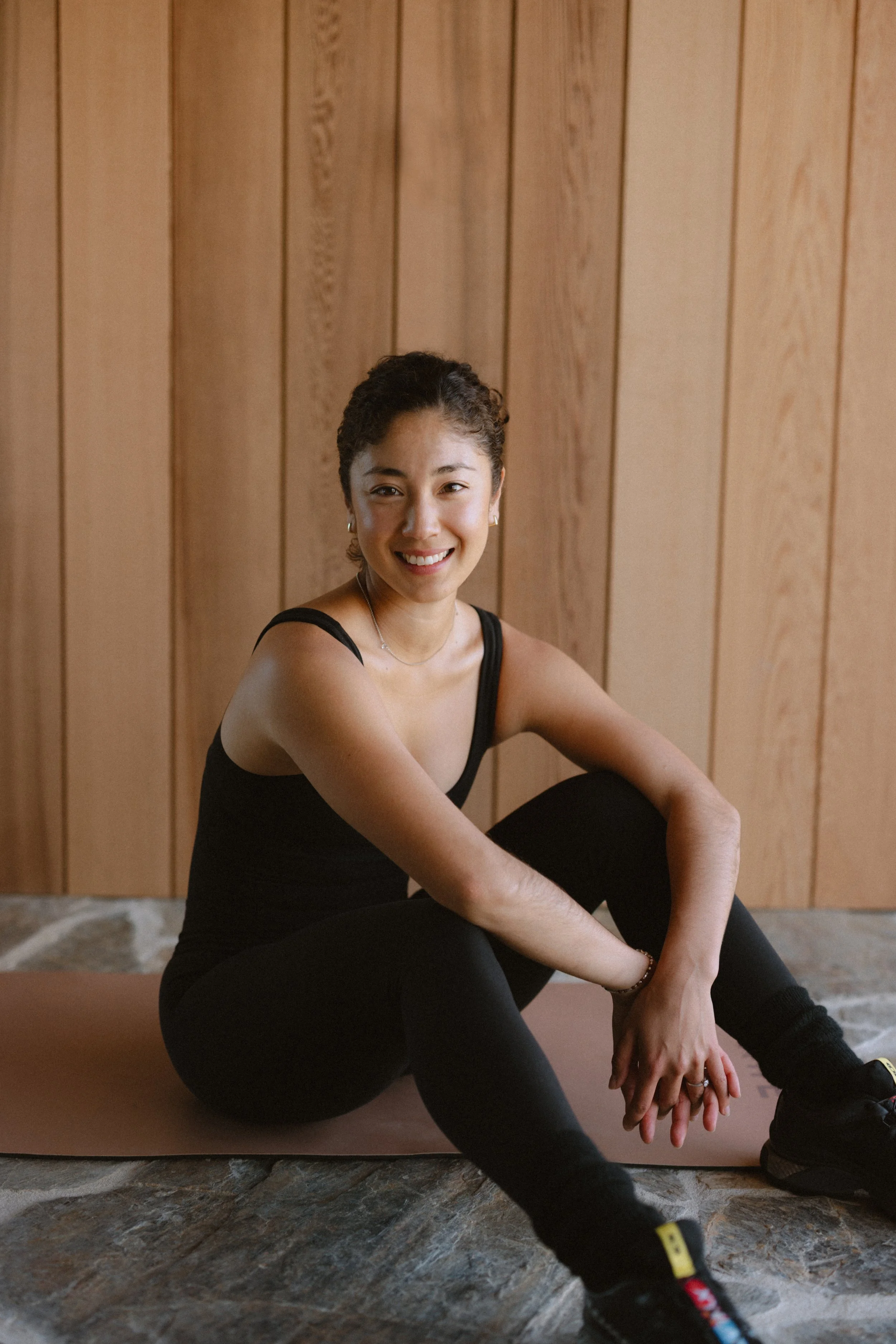 Smiling woman in black athletic wear sitting on yoga mat in front of wooden wall.