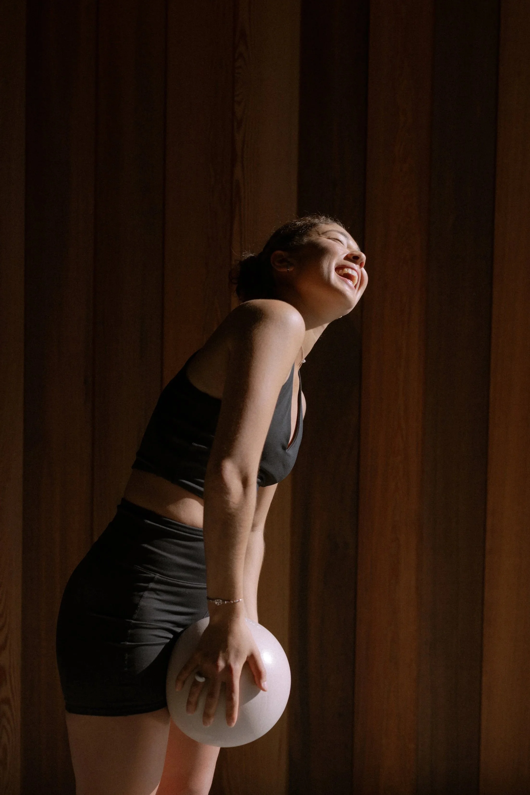 A woman in workout clothing holding a medicine ball, smiling with eyes closed, standing against a wooden wall.