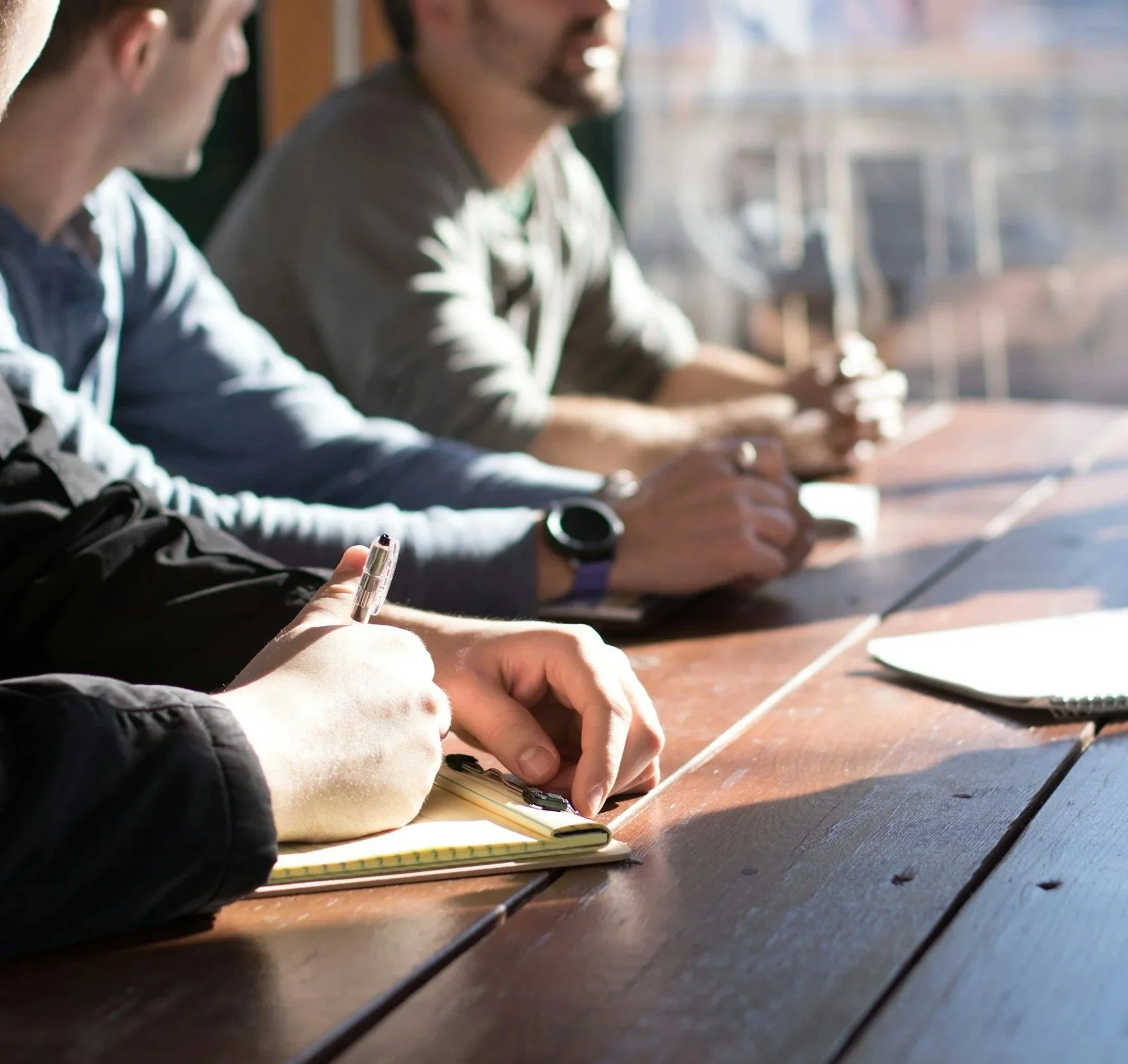 People sitting at a wooden table, writing in notebooks, with sunlight shining through a window in the background.