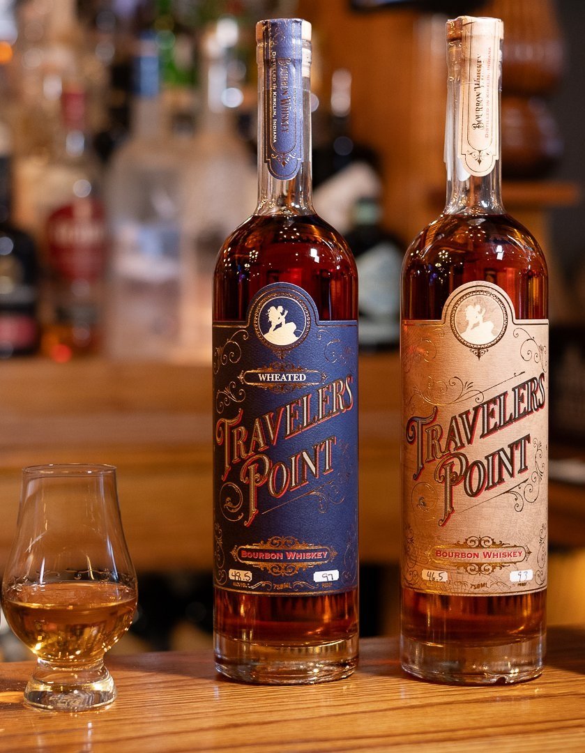 Three bottles of Traveler's Point bourbon whiskey, lined up on a wooden bar counter, with a glass of whiskey beside them. The bottles have textured labels in blue, beige, and maroon colors.