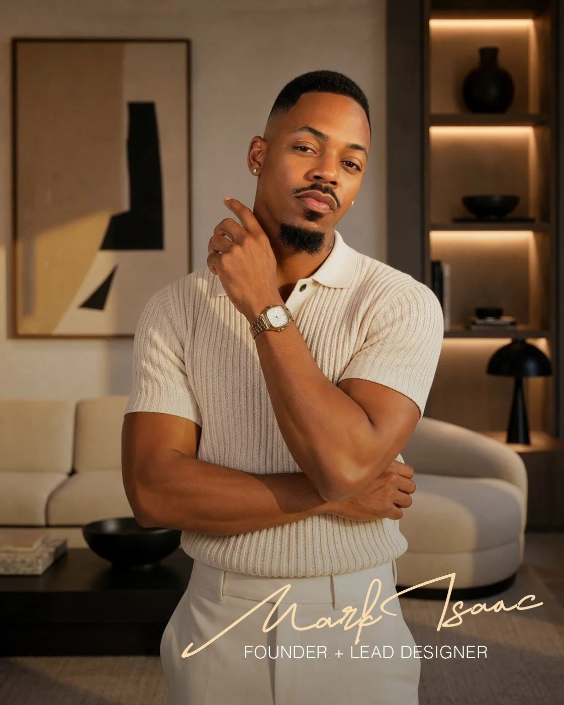 A man with a trimmed beard and short hair wearing a cream-colored, short-sleeve, ribbed polo shirt and white pants, standing confidently with his arms crossed, touching his face with his right hand, in a modern, elegantly decorated room. The background features abstract artwork, a cream-colored sofa, a black sculpture, and built-in shelves with decorative items. The text overlay reads 'Marc Isaac, Founder + Lead Designer.'