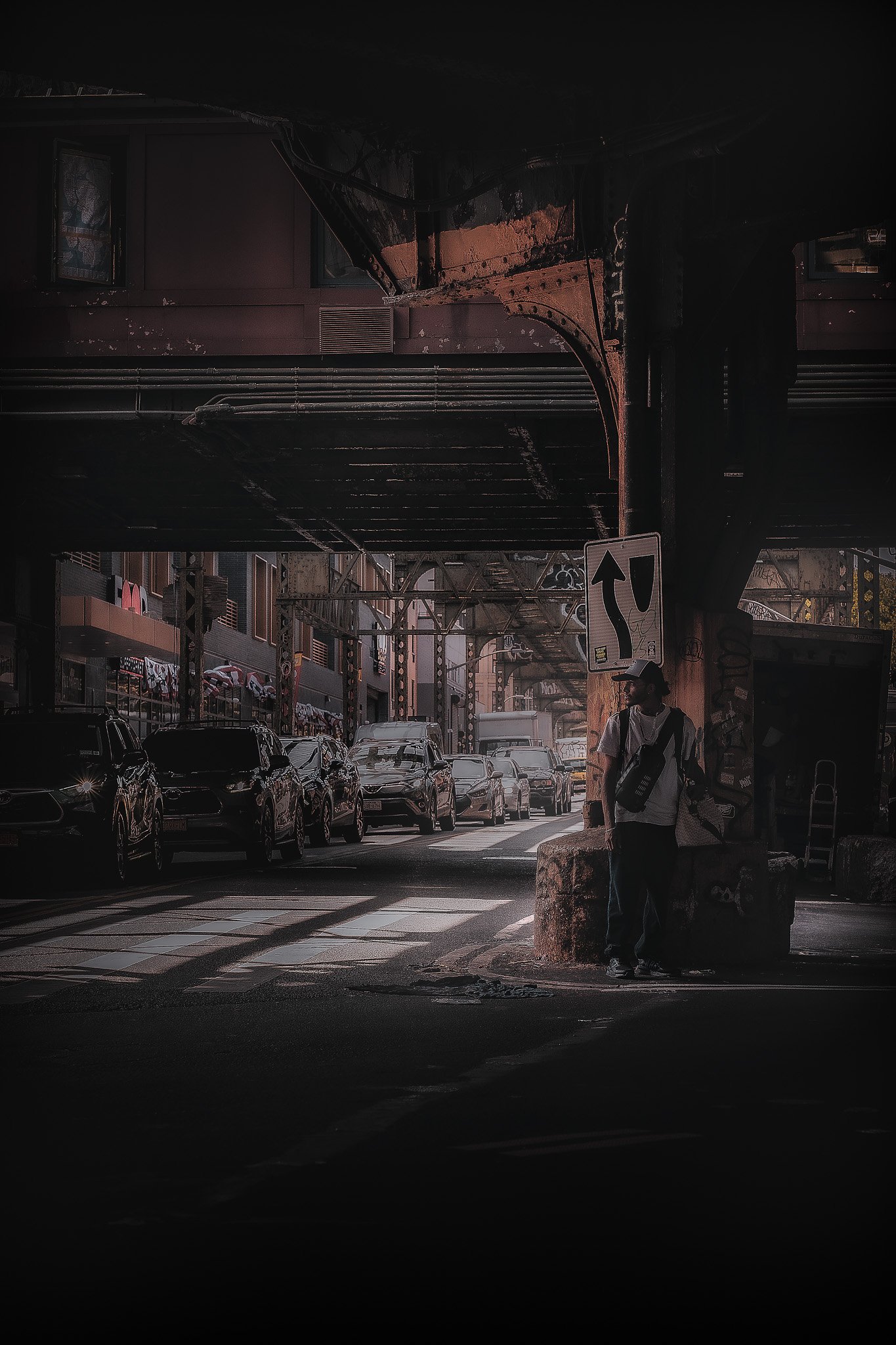 An urban street scene during daytime shows cars parked along the street, with a person standing near a concrete post holding a bag. Overhead train tracks and a sign indicating a left or straight direction are visible above. Buildings and graffiti can be seen in the background.