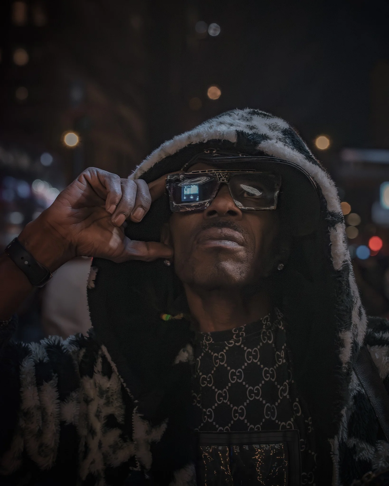 A person wearing a fur-lined hooded jacket and dark sunglasses, with their hand adjusting the sunglasses, standing outdoors at night with blurry city lights in the background.