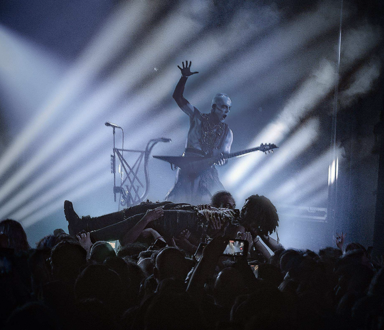 Live concert scene with a guitarist on stage, illuminated by beams of light; a performer is being lifted by the audience, with a crowd in the foreground.