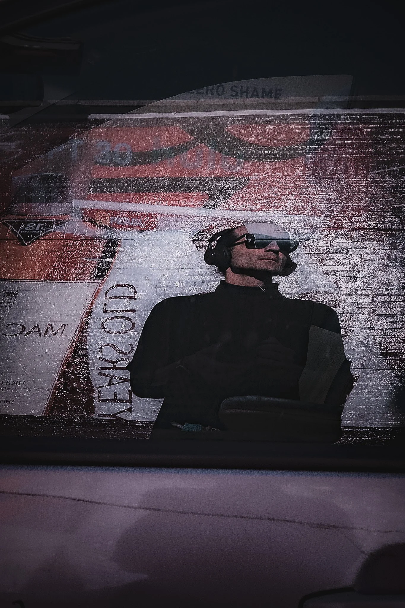 Reflected image of Mac wearing sunglasses and a headset, visible through a rain-covered car window with some signage and text visible.