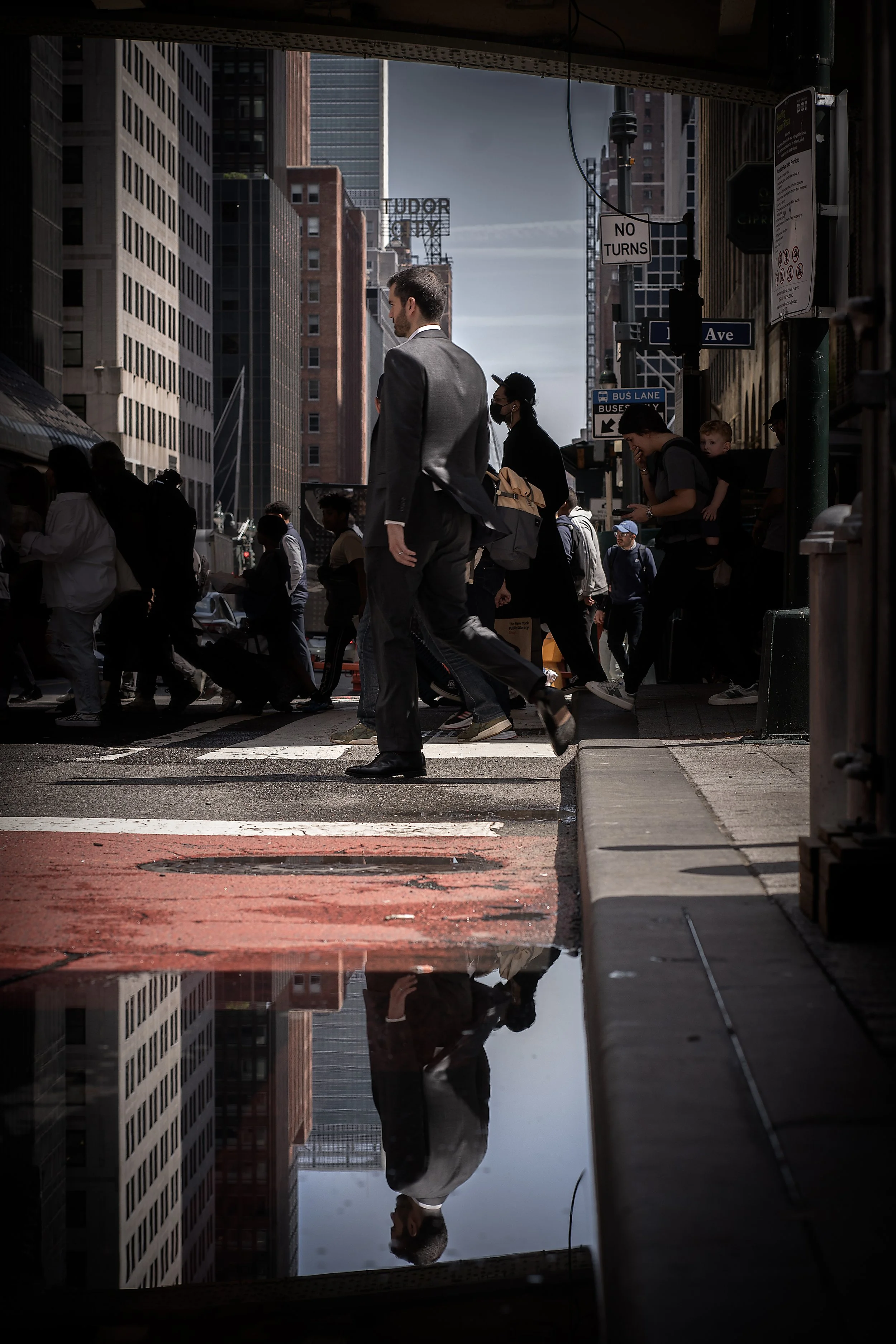 Urban street scene with people crossing a crosswalk, tall buildings, a puddle reflecting the scene, and signs including 'No Turns' and 'Ave'.