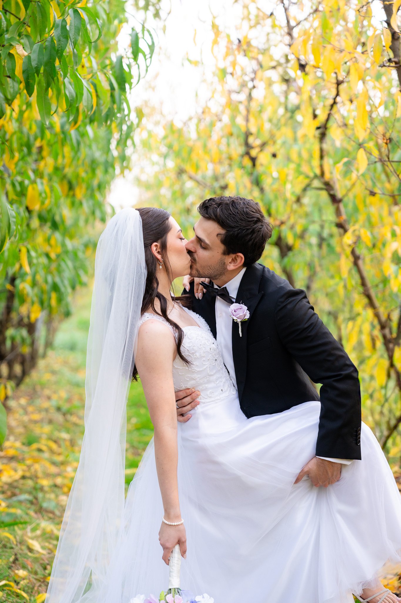 A bride and groom kissing outdoors during autumn, surrounded by trees with yellow leaves. The groom lifts the bride in a wedding pose, and she holds a bouquet on the ground.