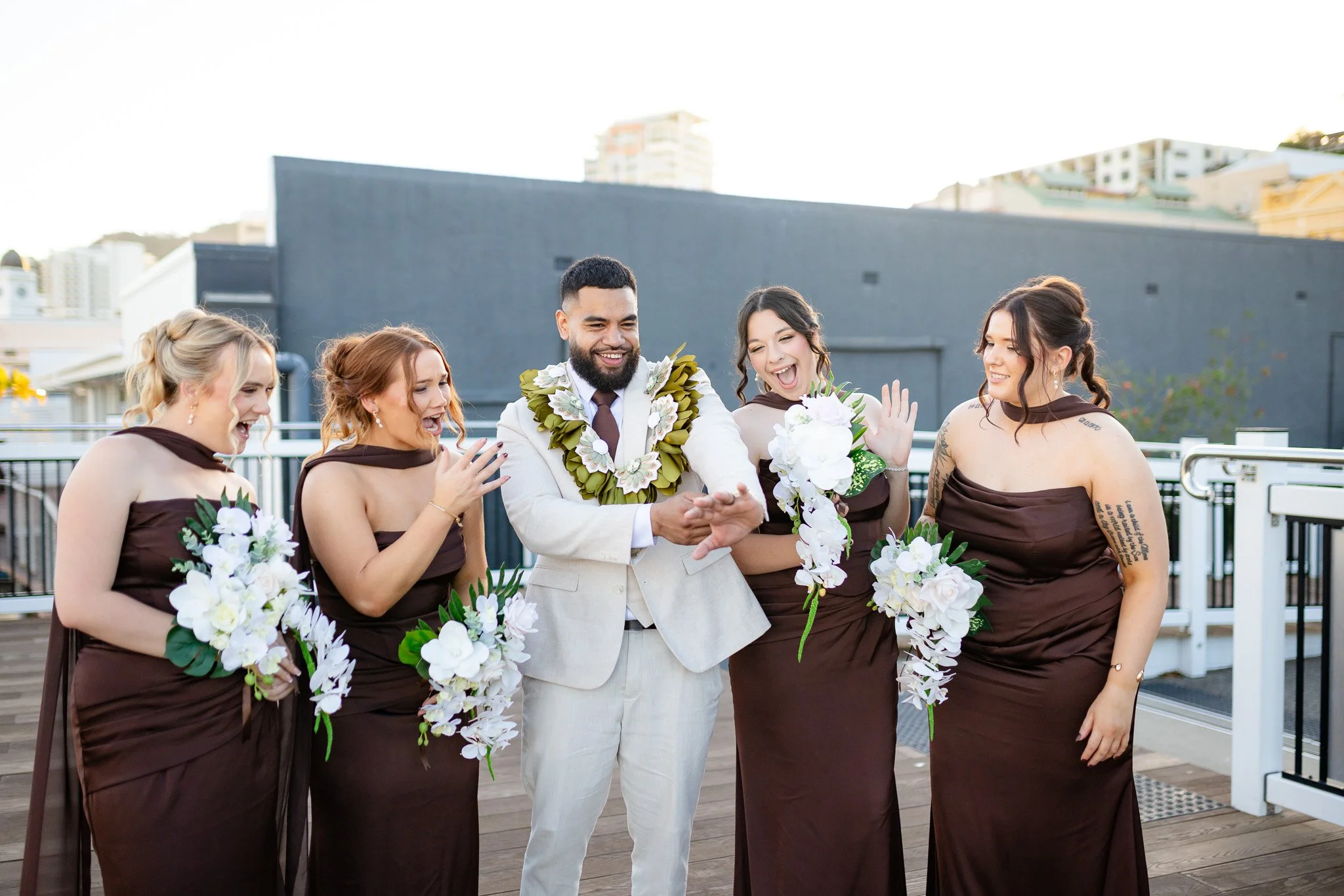 A group of five people, four women and one man, dressed in formal attire, are standing together outdoors, celebrating and smiling, with some holding floral bouquets.