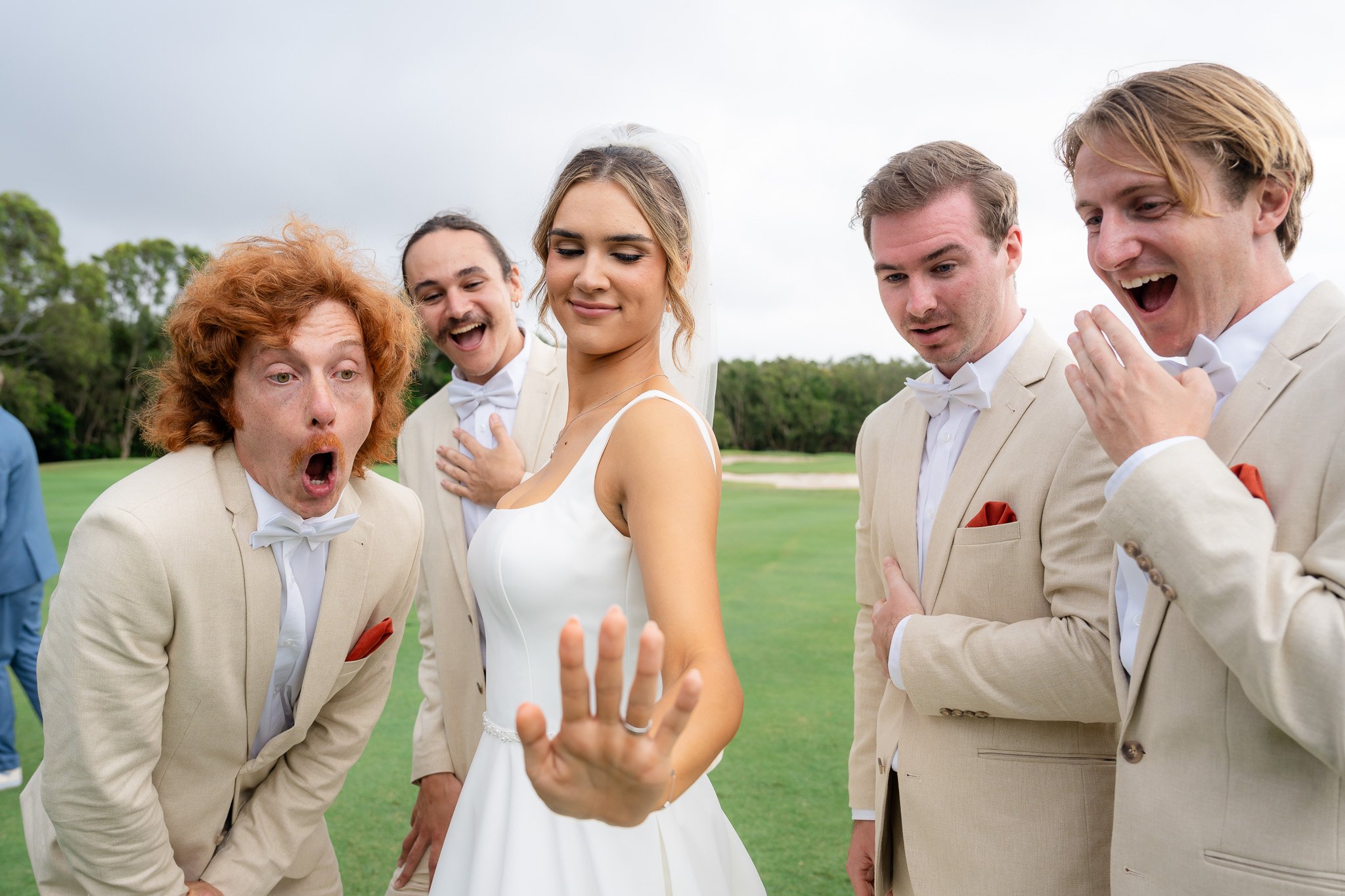 Group of five people dressed in formal attire at an outdoor wedding, with a woman in a wedding dress, and four men in cream-colored suits, some with surprised expressions, one with a hand on his chest and one smiling broadly.