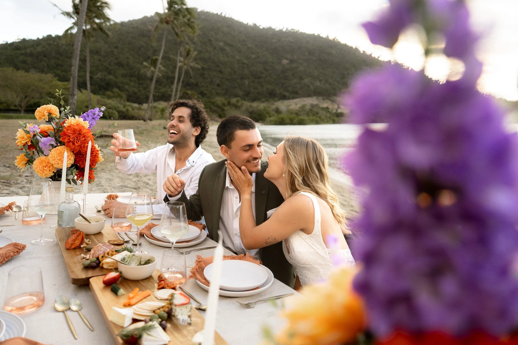 Group of four people at a dinner party on a beach, with a scenic mountain background, decorated table with flowers, various dishes, and drinks, enjoying conversation and laughter.