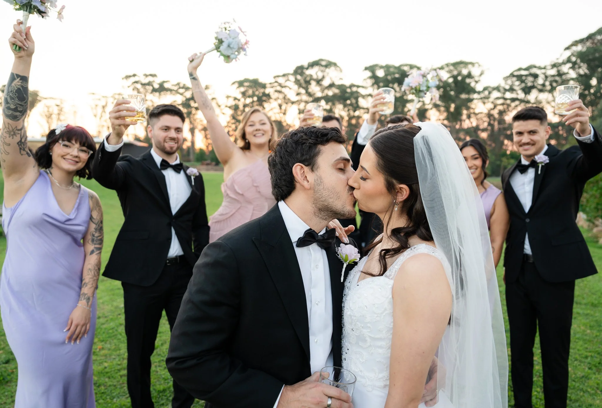 A wedding celebration outdoors at sunset with a bride and groom kissing, surrounded by friends in formal attire raising glasses and holding bouquets.