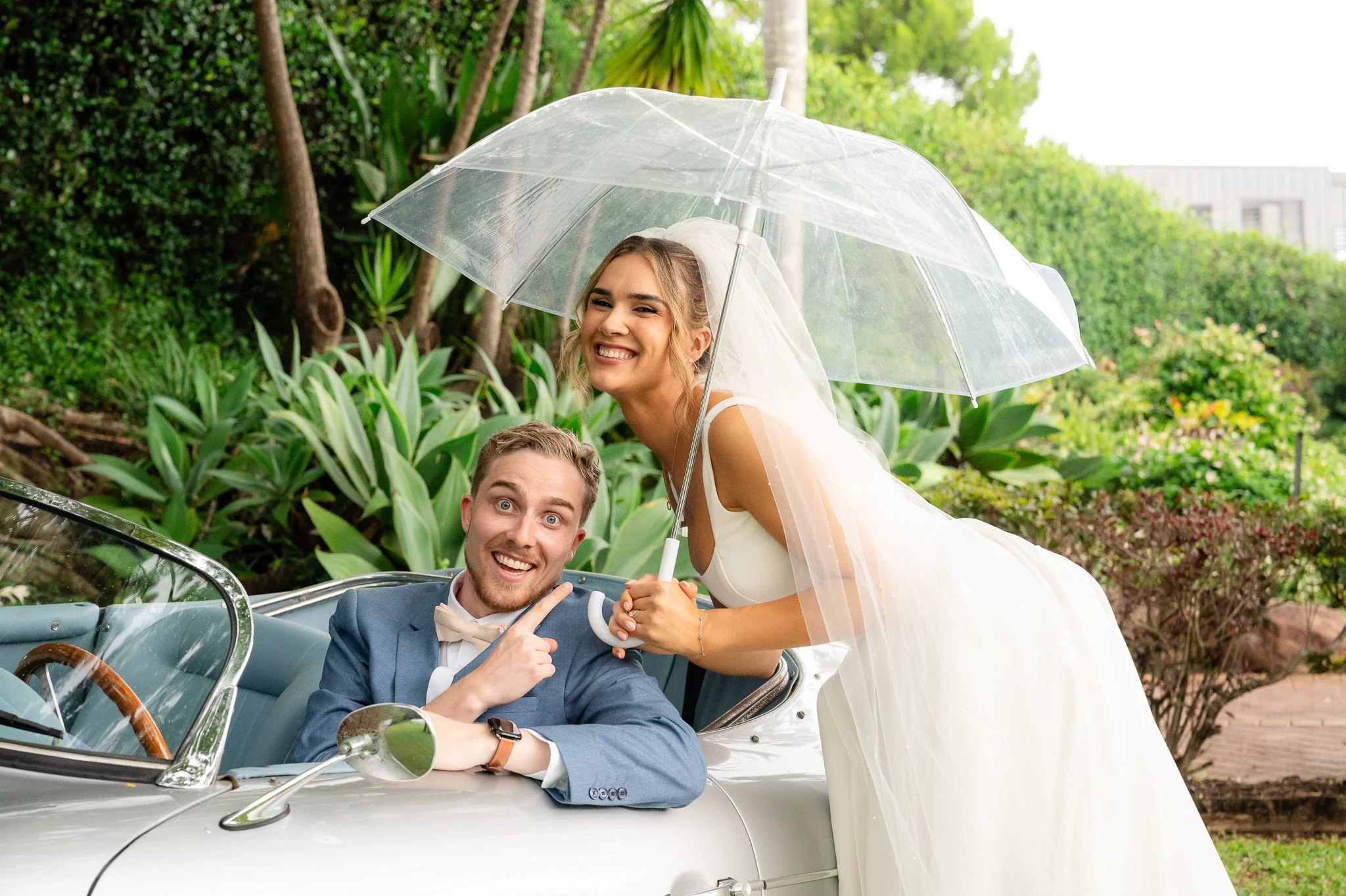 A smiling bride and groom inside a vintage convertible car, with the bride holding a transparent umbrella, surrounded by lush greenery.