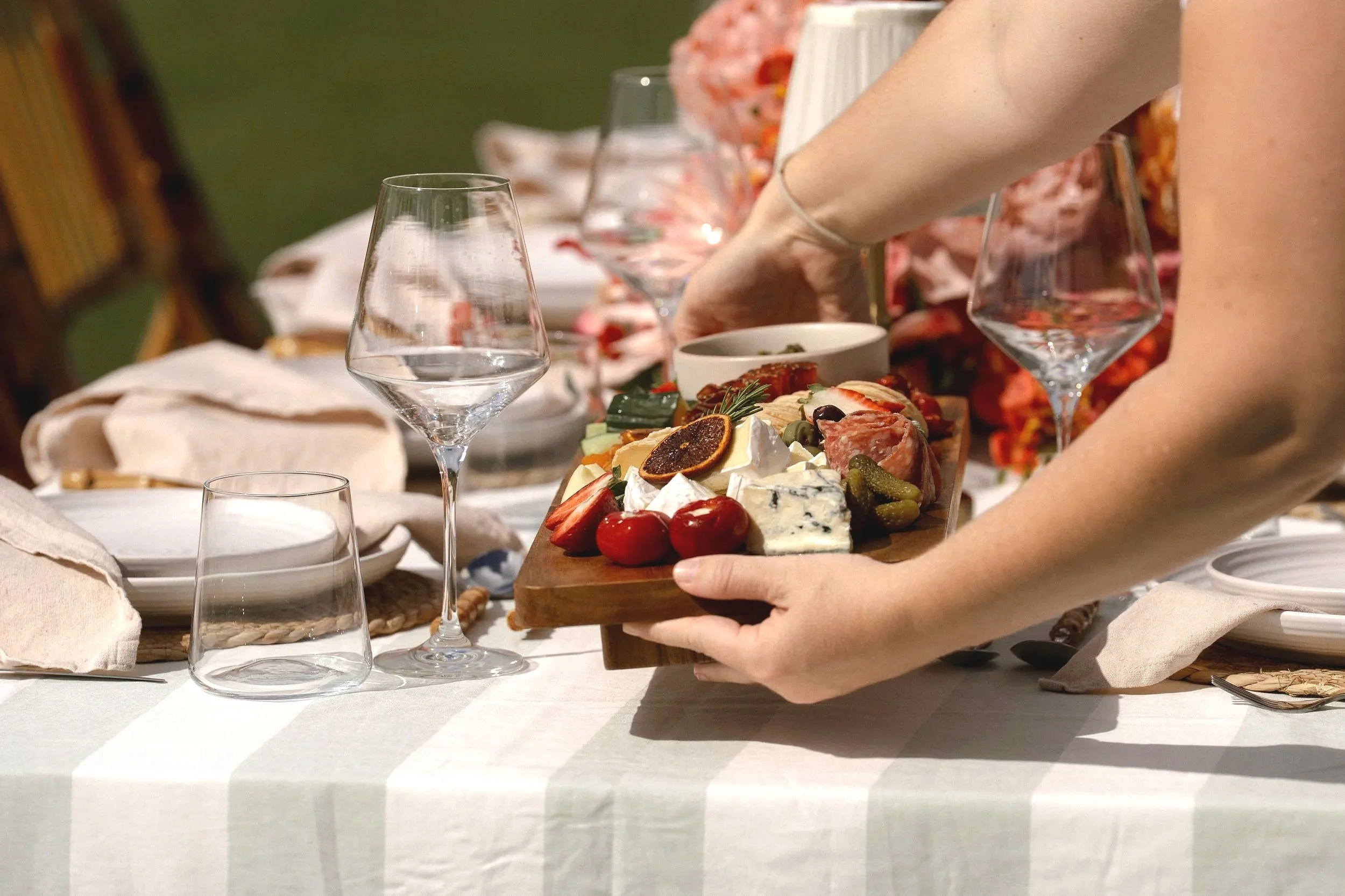 Person holding a wooden cheese board with various cheeses, strawberries, and herbs on a dining table set for a meal.