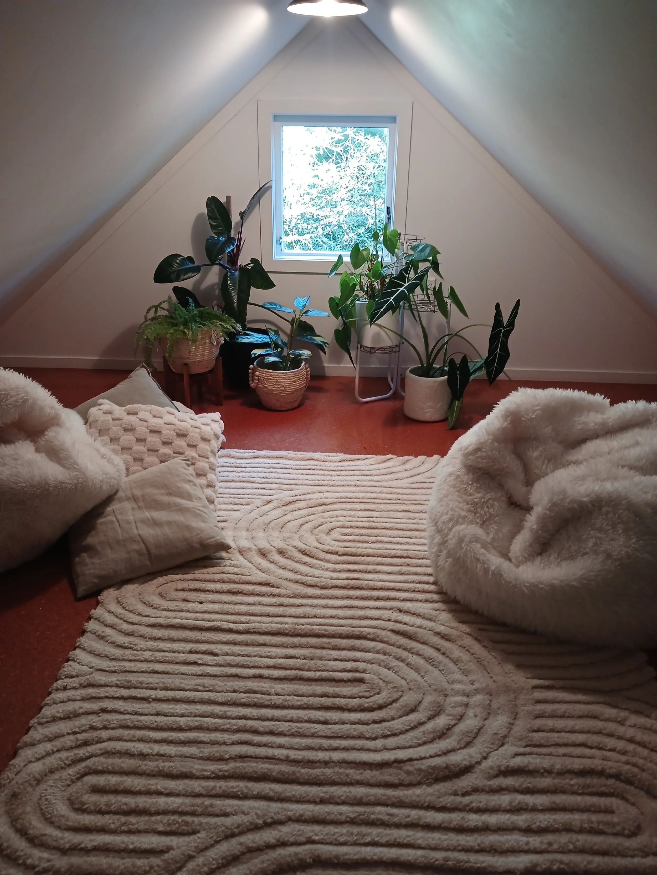 Cozy attic room with a sloped ceiling, small window, large green potted plants, pillows, and a plush beige rug.