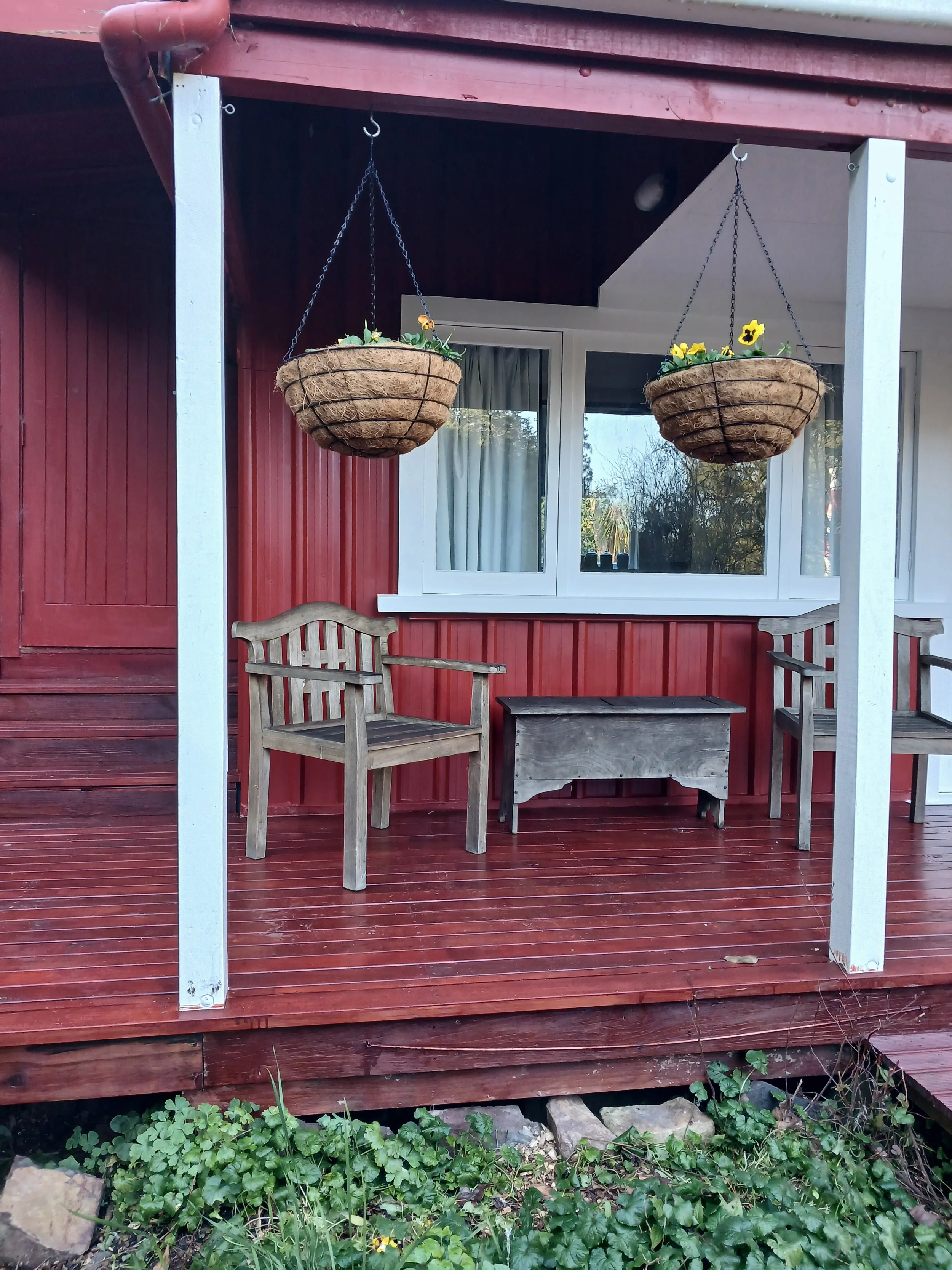 A porch with two wooden chairs, a small wooden table, and two hanging flower baskets containing yellow flowers. The porch has a red painted wooden floor and walls, with a large window behind the chairs.