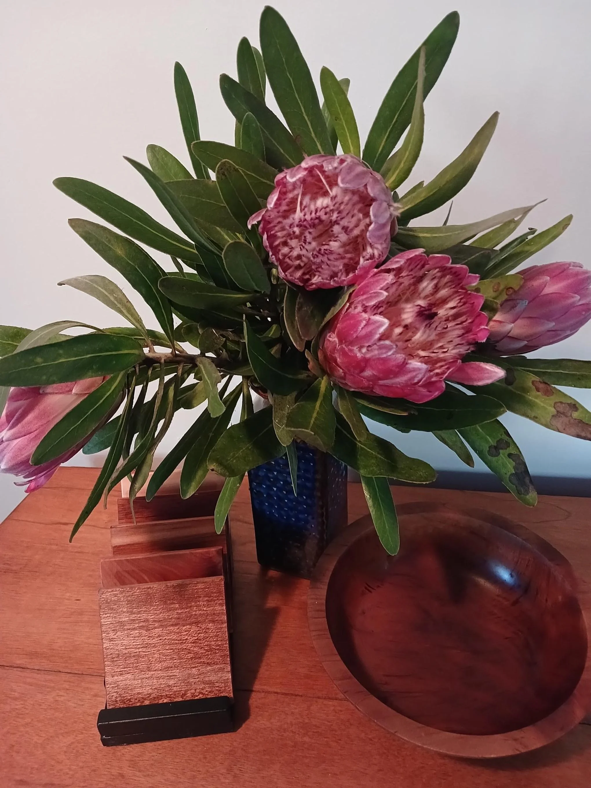 A bouquet of pink protea flowers with green leaves in a dark blue textured vase on a wooden table, with a wooden bowl next to it and a small stack of wooden blocks.