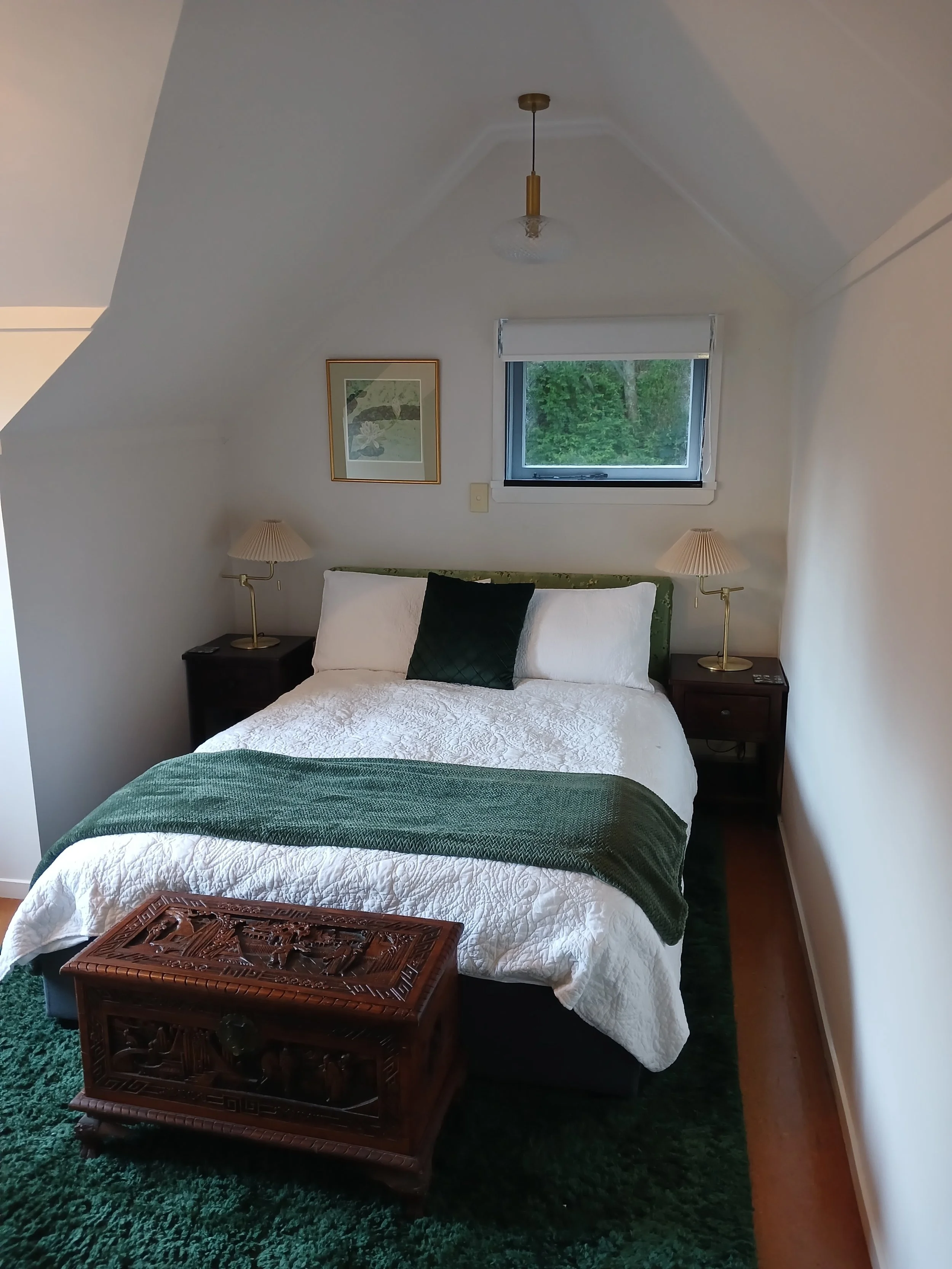 A cozy bedroom with a double bed, white bedding, a dark green throw blanket, and a black accent pillow. There are two nightstands with lamps on each side and a small window with greenery outside. An ornate wooden chest sits at the foot of the bed. Th