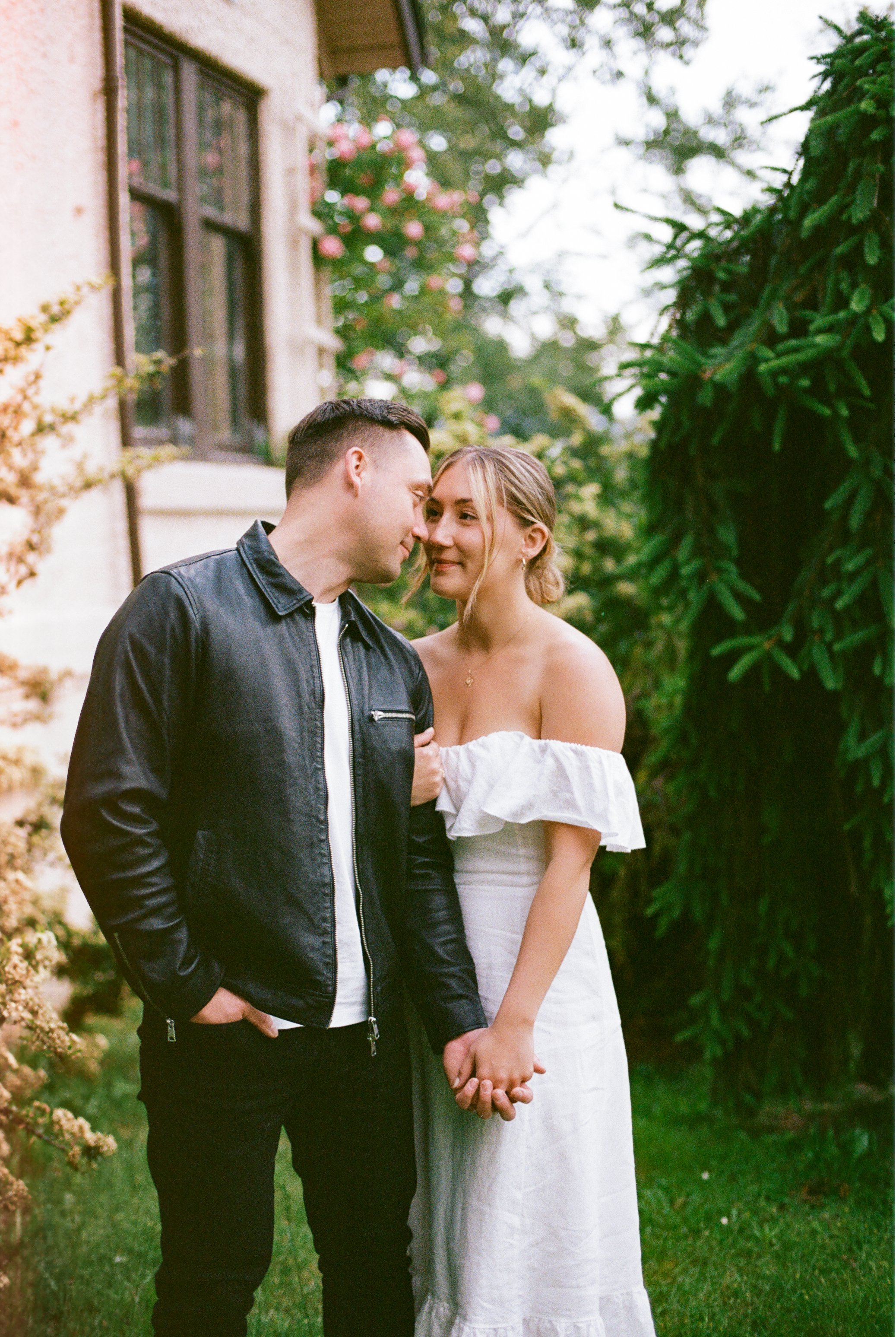 A young couple stands close together outdoors, holding hands, sharing an intimate moment, with a house, outdoor greenery, and pink flowers in the background.