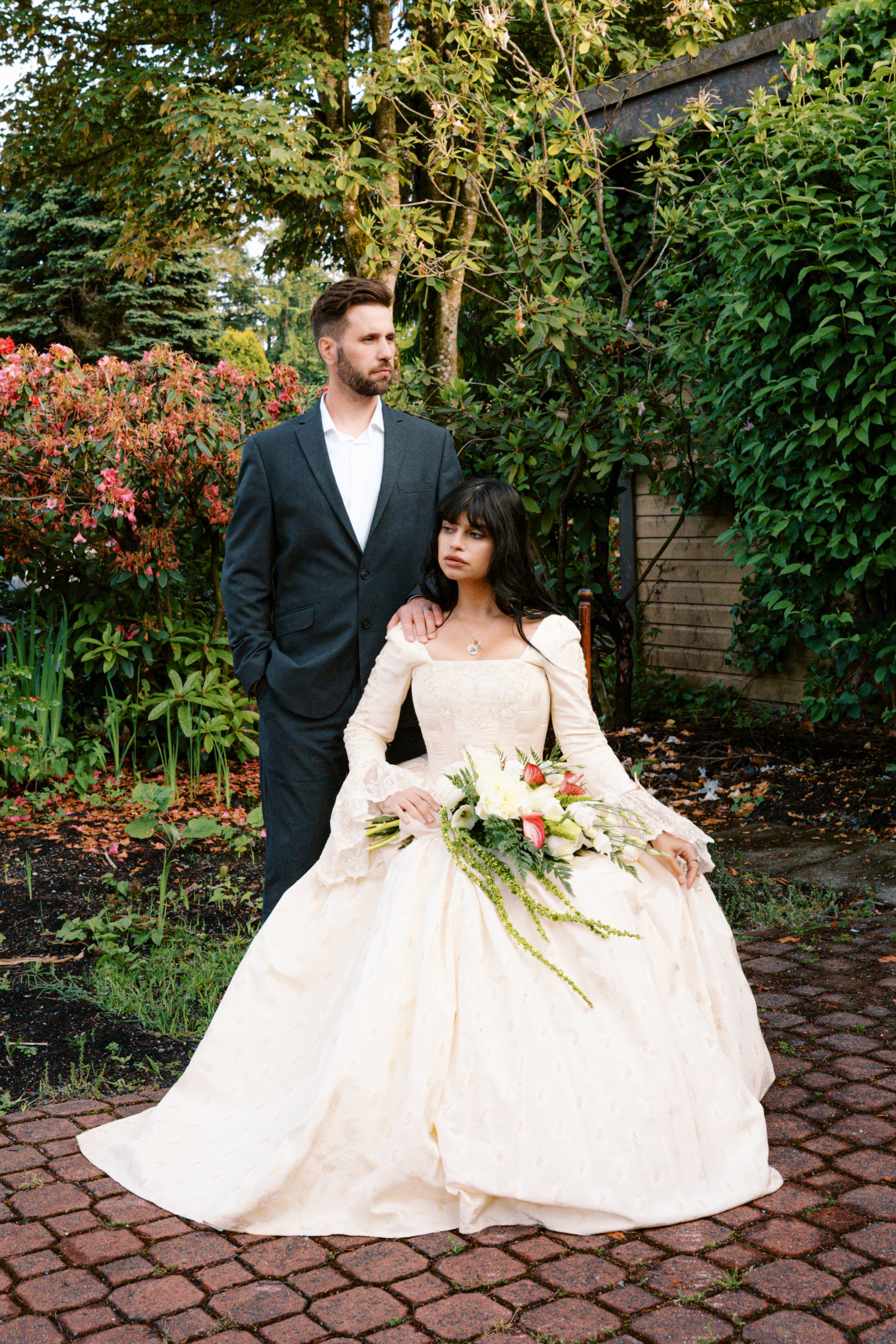 A man in a dark suit and a woman in a vintage wedding dress holding a bouquet of white and pink flowers pose outdoors on a brick pathway surrounded by greenery.