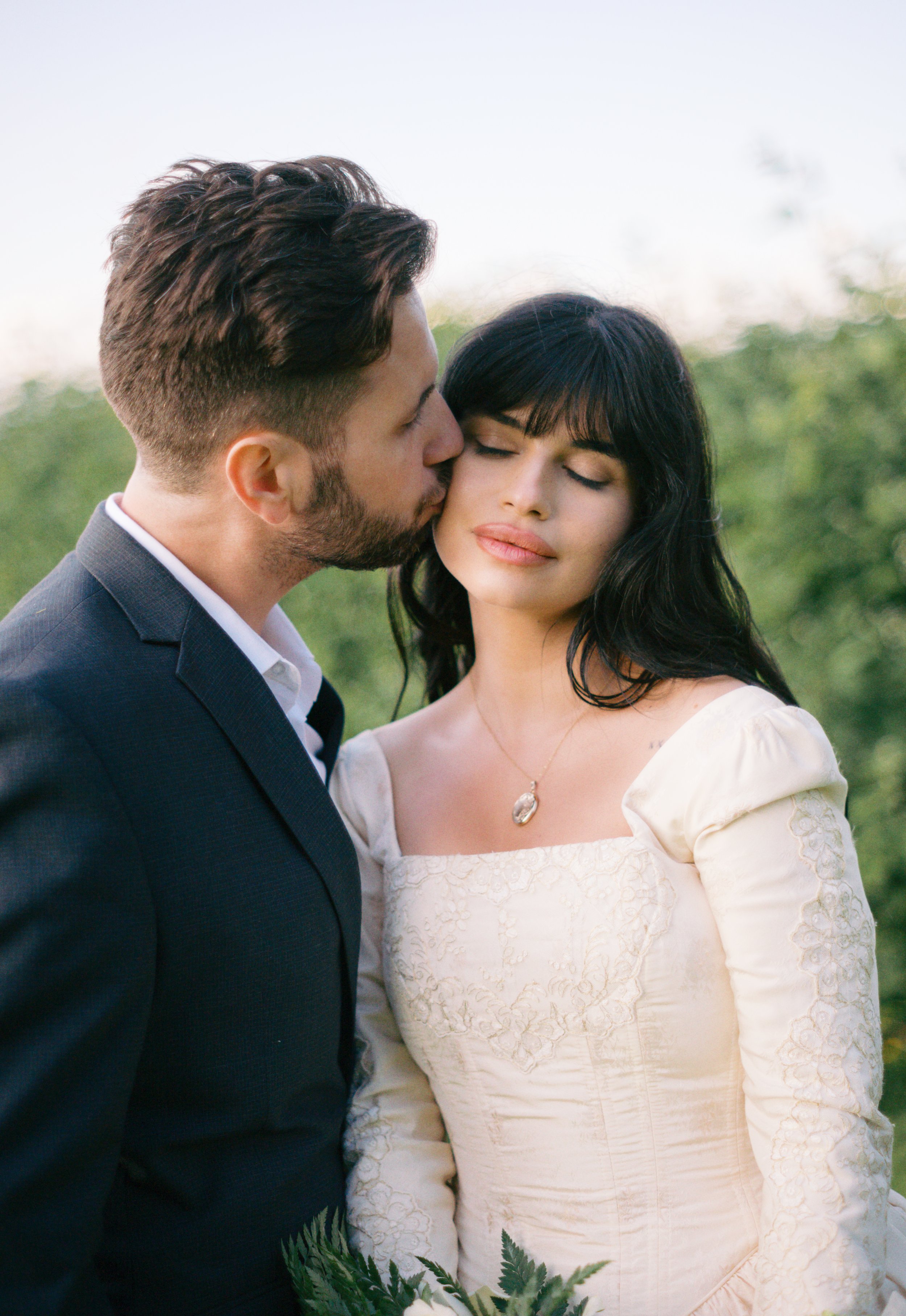 A man in a dark suit kisses a woman in an off-white dress with dark hair and a pendant necklace outdoors with greenery in the background.
