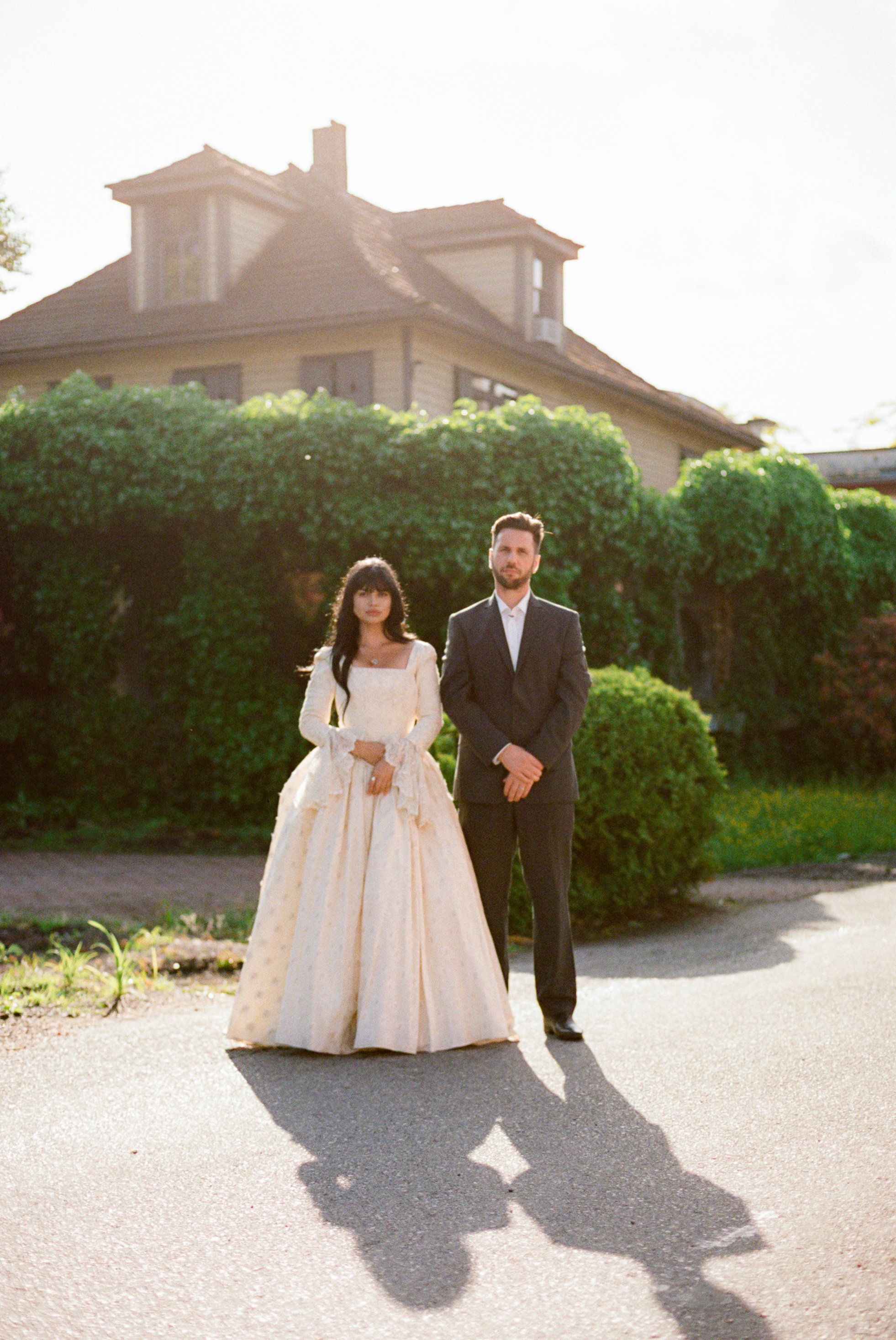 A woman in a vintage wedding gown and a man in a dark suit are standing outdoors on a paved surface in front of green bushes and a house, with sunlight casting long shadows.