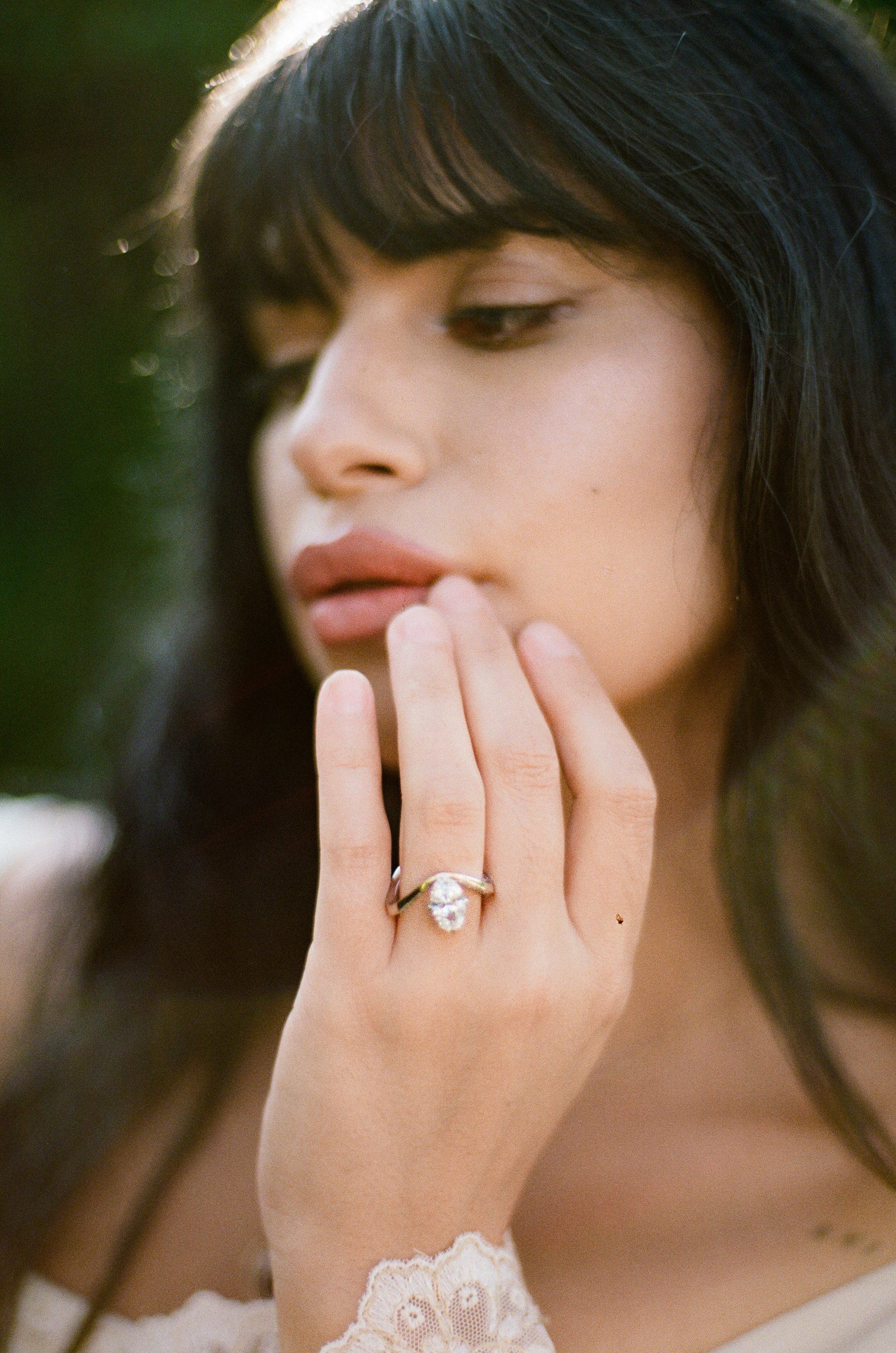 A woman with dark hair and light skin gently touching her lips with her hand, showcasing a diamond ring on her finger.