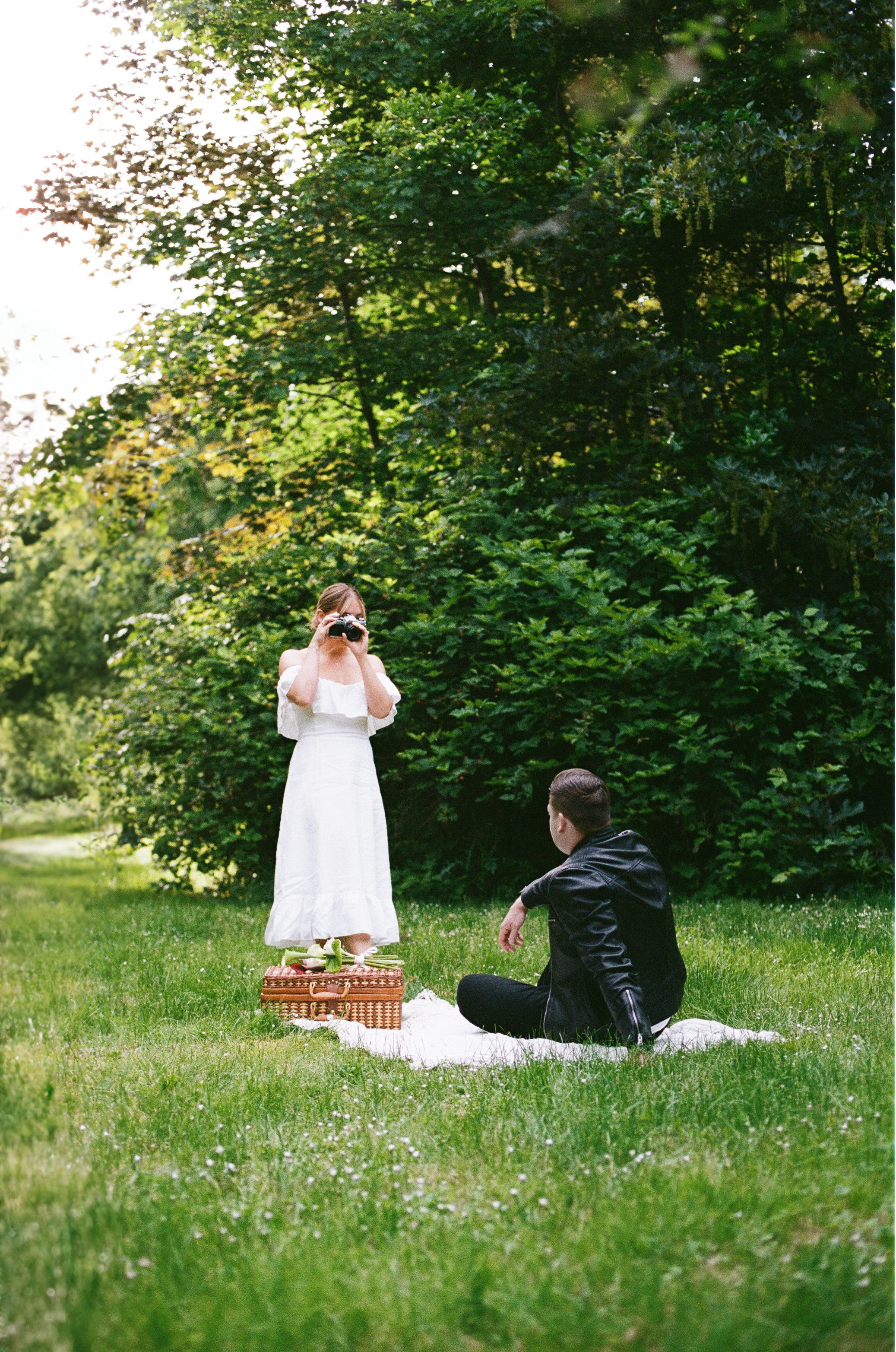 A woman in a white dress takes a photograph of a man sitting on a blanket on the grass during a picnic outdoors, surrounded by lush green trees.