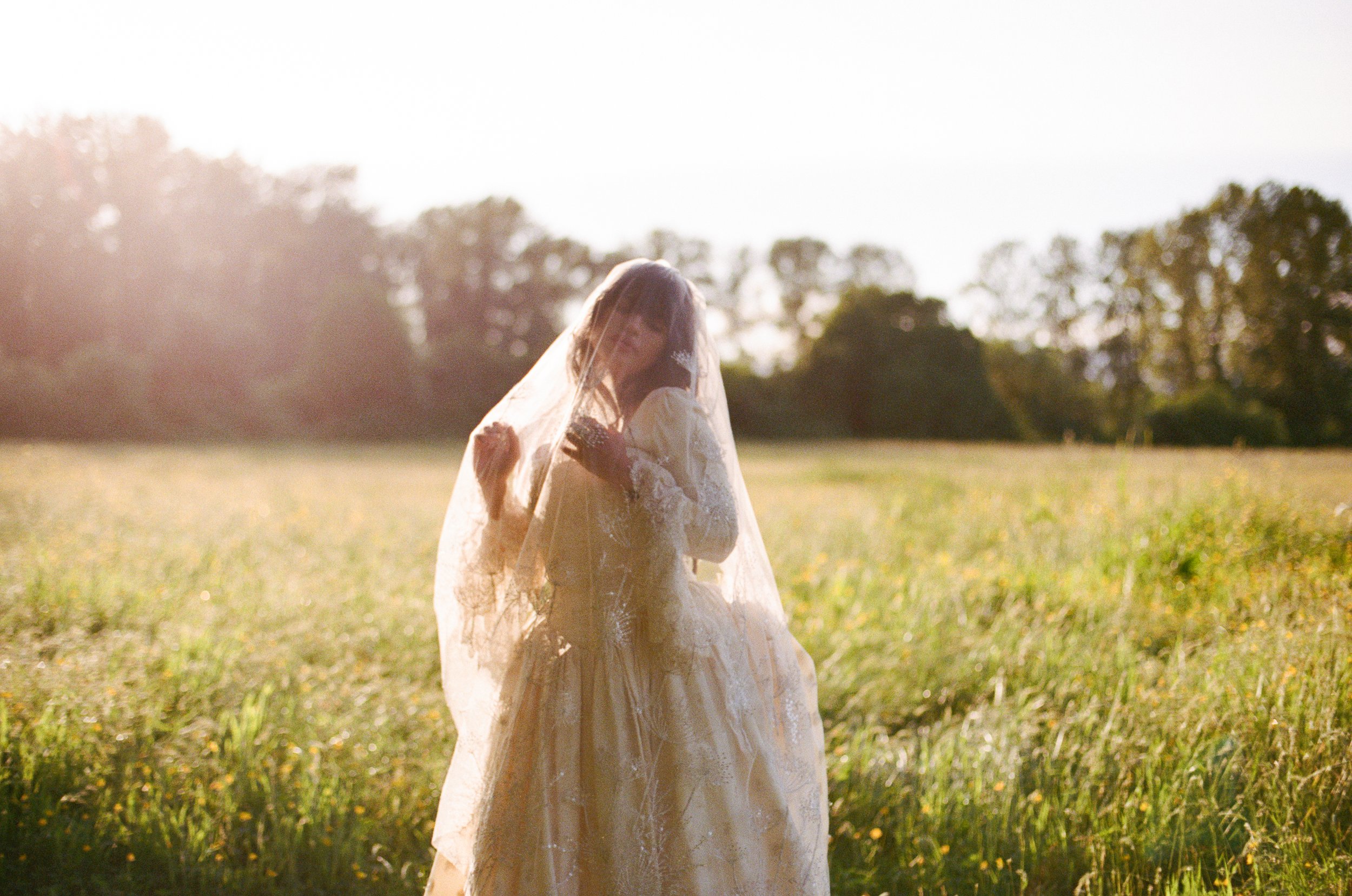 A bride standing in a sunlit grassy field, wearing a wedding dress and veil, with trees in the background.