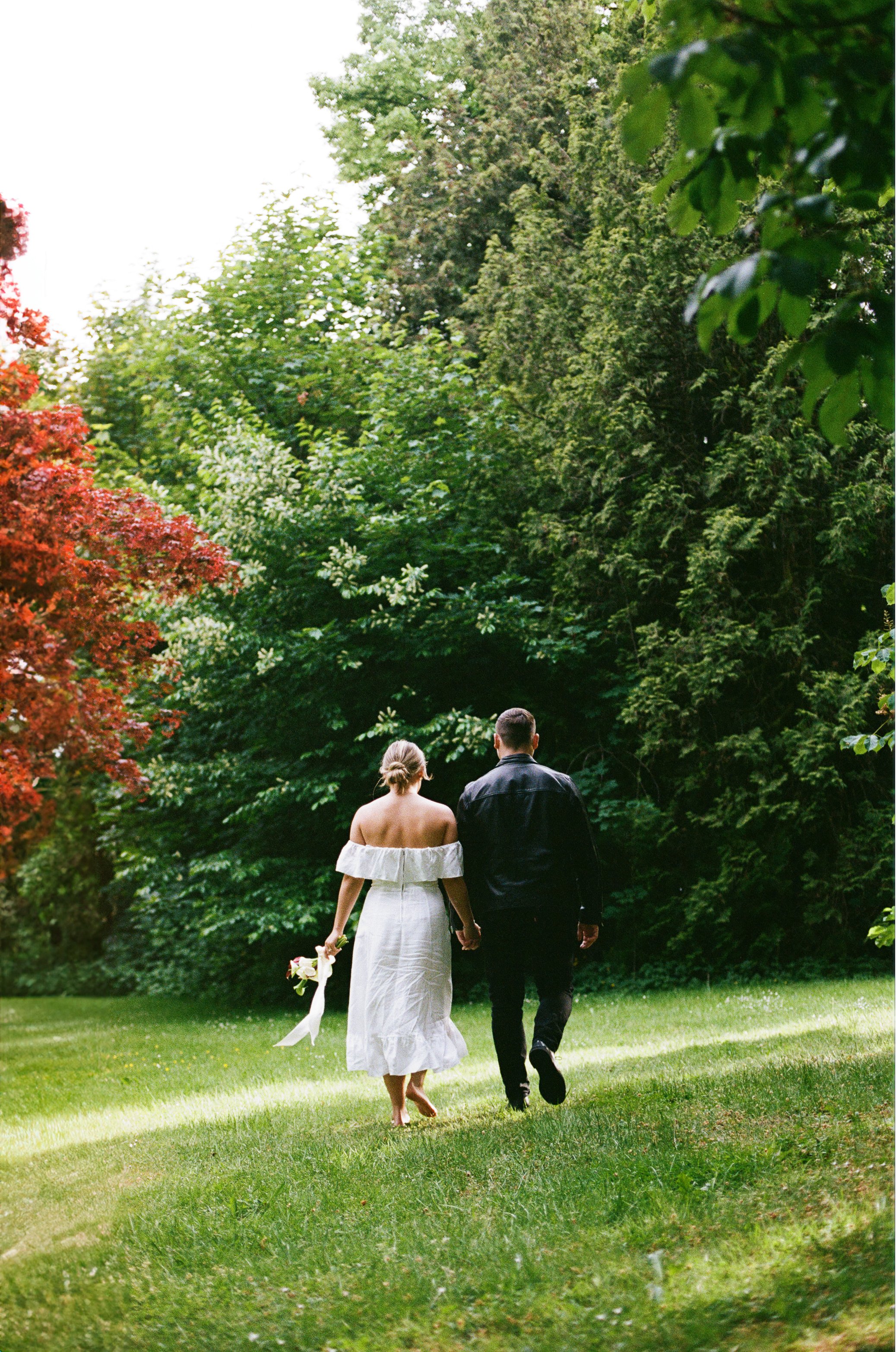 A couple walking hand-in-hand through a grassy park surrounded by lush green trees, with the woman in a white off-shoulder dress holding a bouquet.