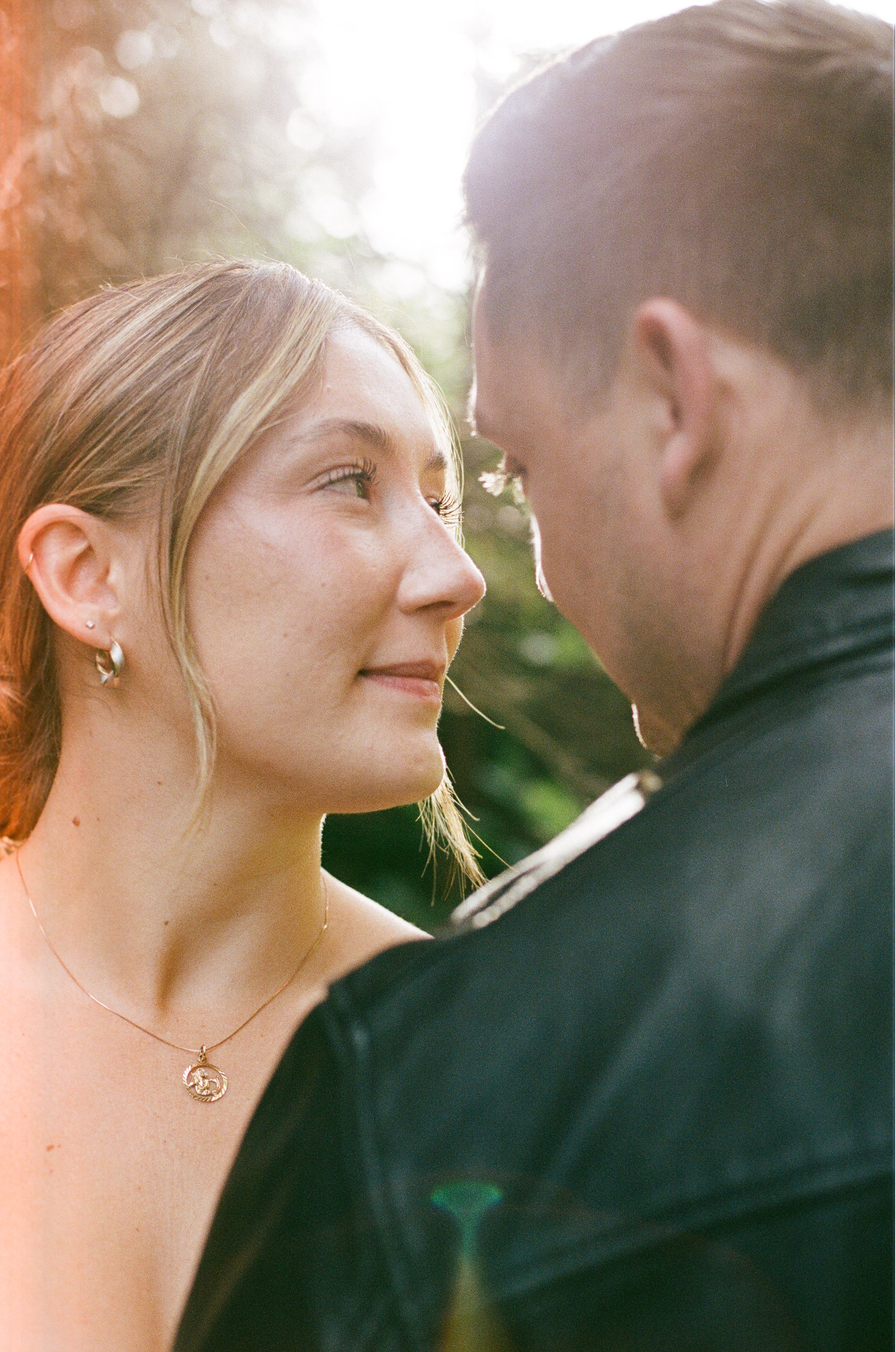 A woman and man facing each other closely outdoors, the woman smiling softly, sunlight filtering through trees in the background.