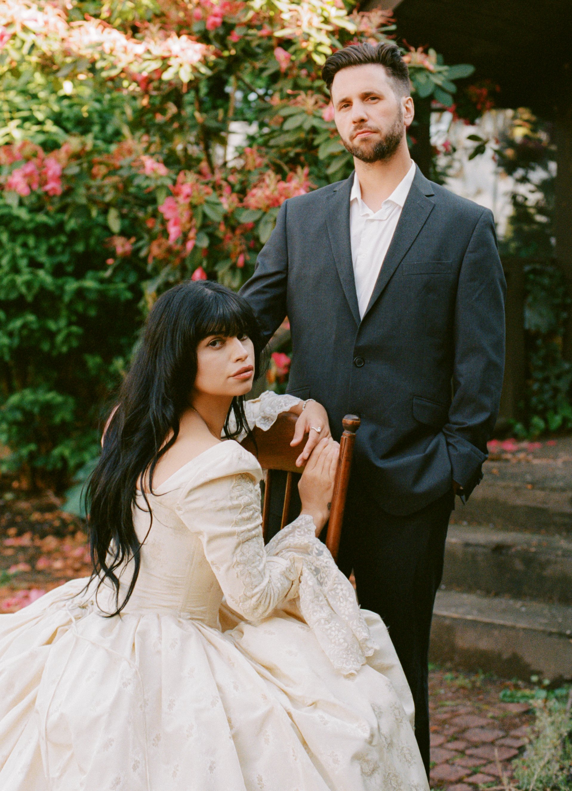 A woman in a vintage cream-colored gown with lace details and long black hair is seated on a wooden chair, holding onto its backrest. A man in a dark suit and white shirt stands beside her, resting his hand on her shoulder. They are outdoors in a gar