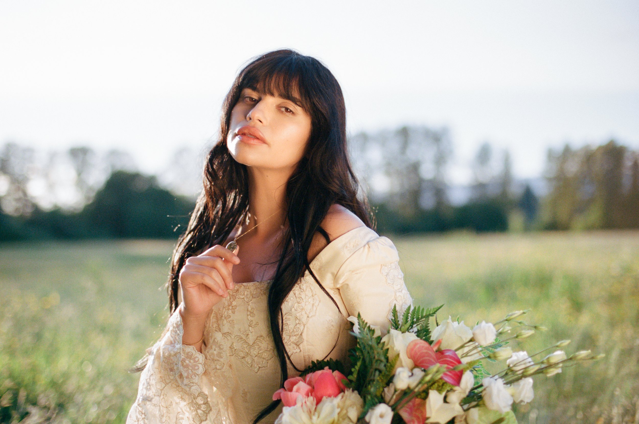 A woman with long dark hair and light skin holding a bouquet of pink and white flowers in an outdoor field during daytime.
