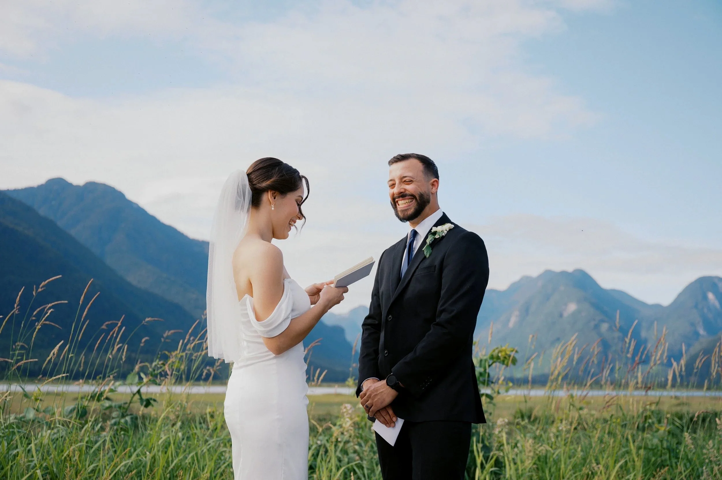 A bride and groom standing outdoors in a field with mountains in the background, exchanging vows and smiling during their wedding ceremony.