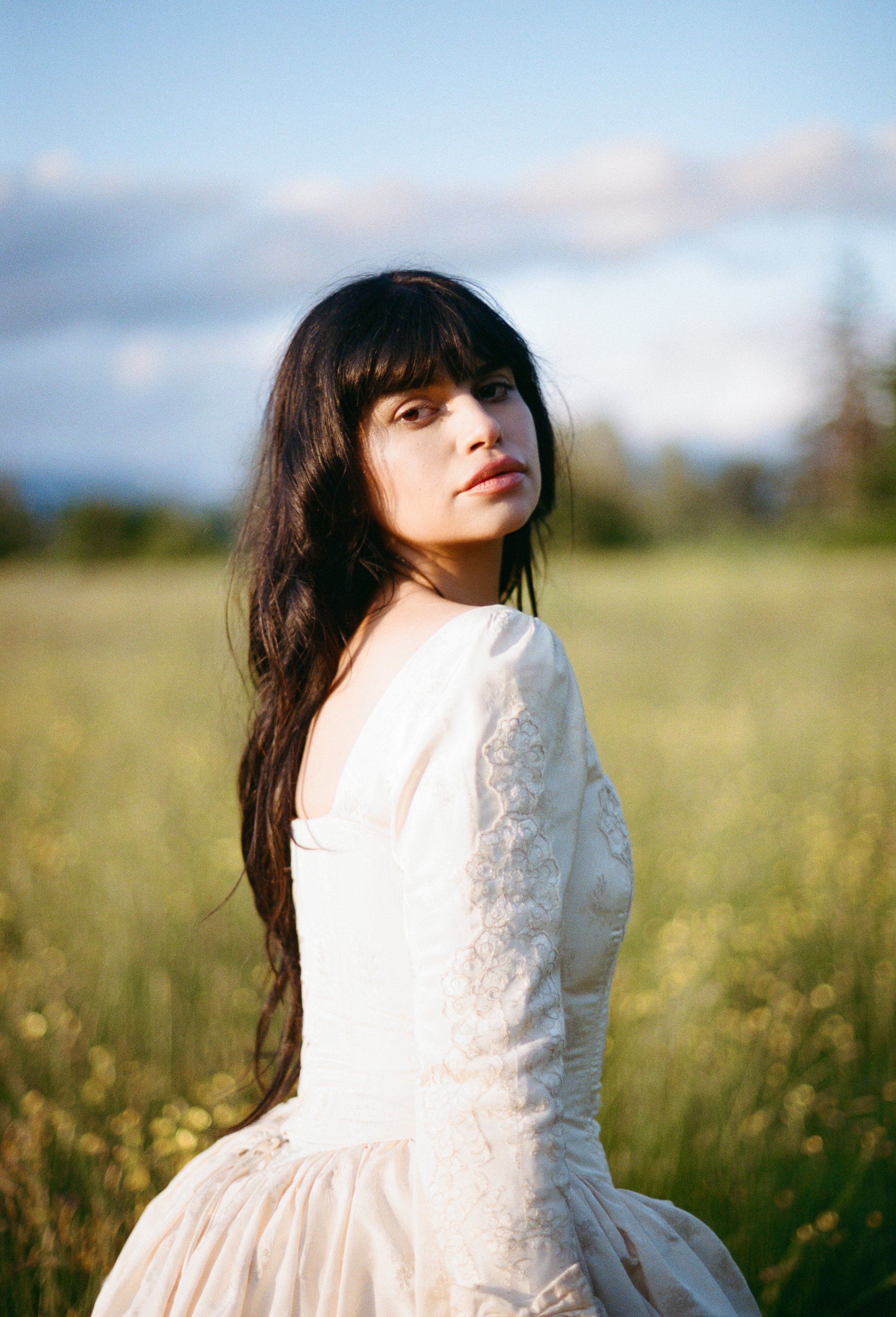 A woman with long dark hair and bangs wearing a vintage cream-colored dress sitting in a grassy field with blue sky and clouds in the background.