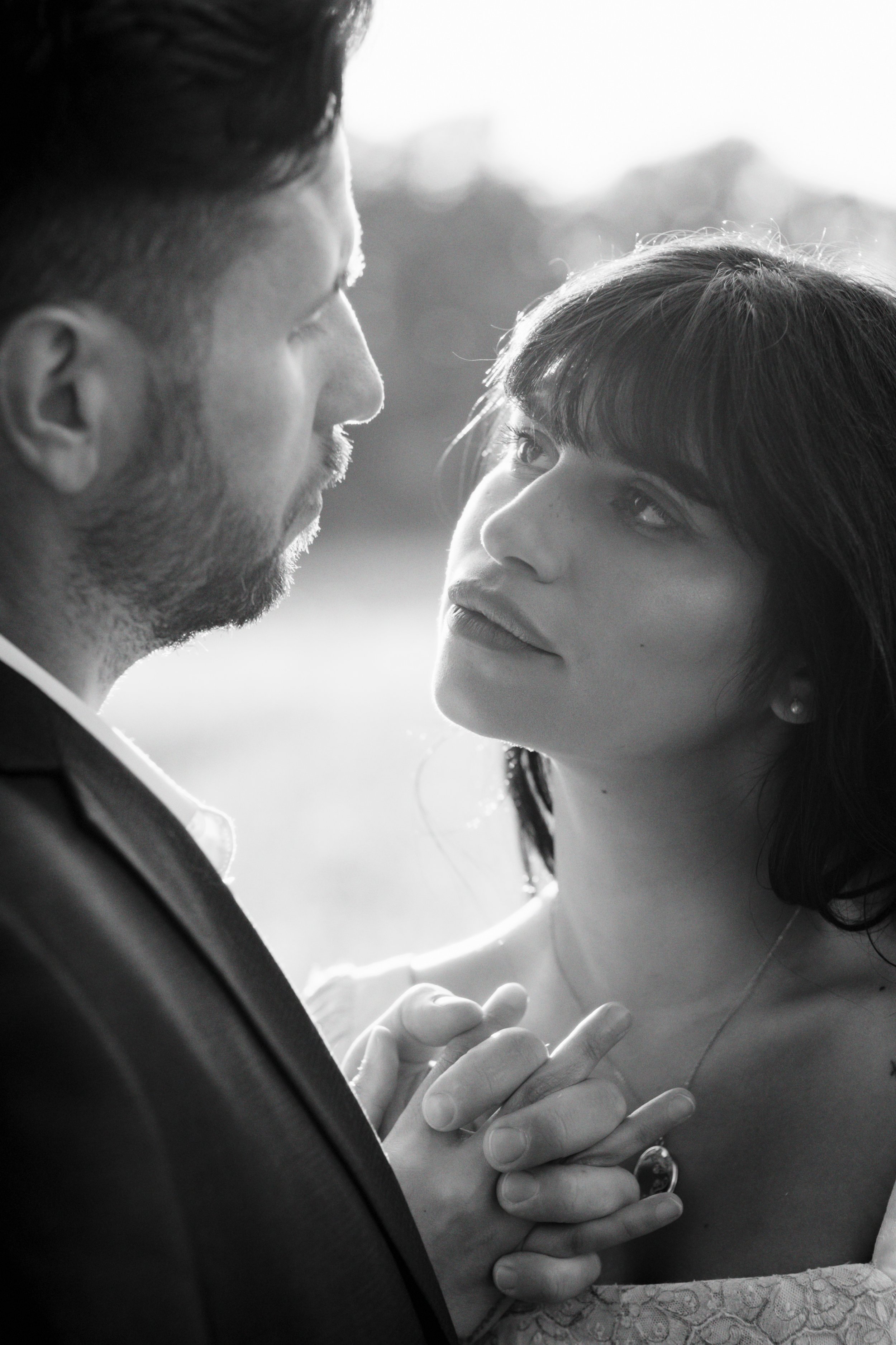 A black and white photo of a couple holding hands and looking into each other's eyes, close-up of their faces with a blurred outdoor background.